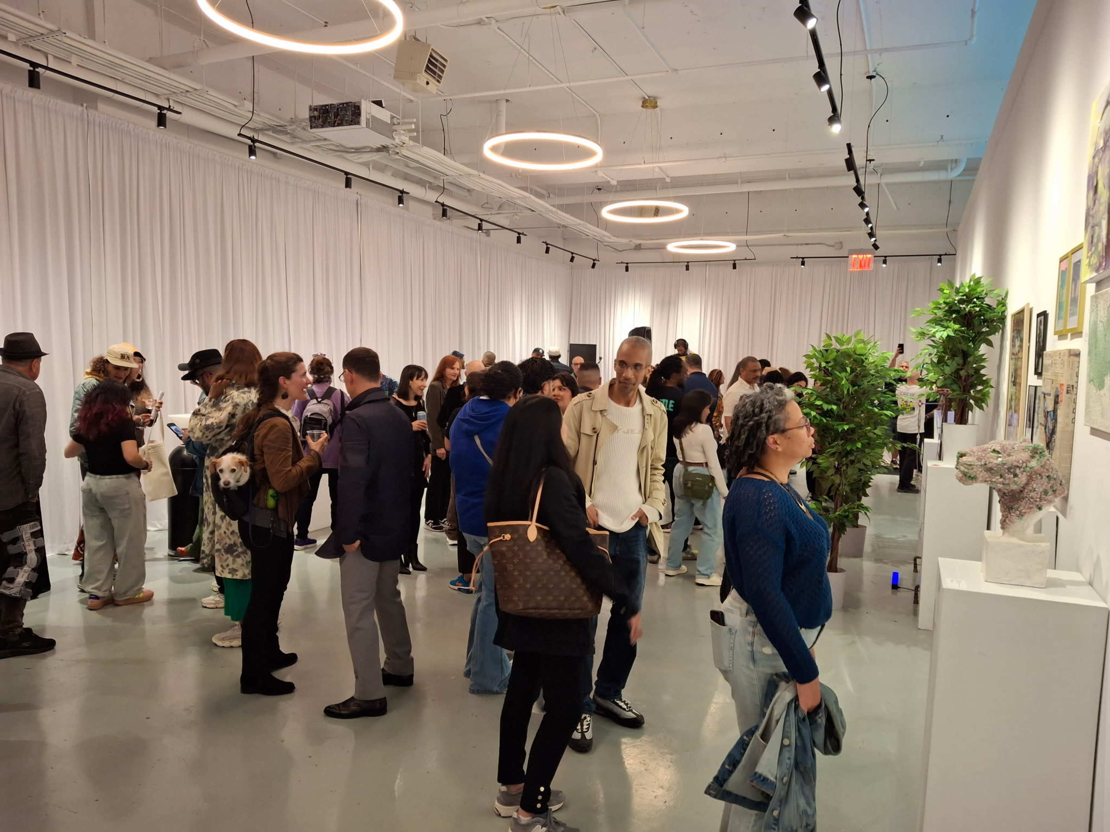 A crowd of people is gathered in an exhibition space with white walls and hanging lights, examining various artworks displayed on stands.