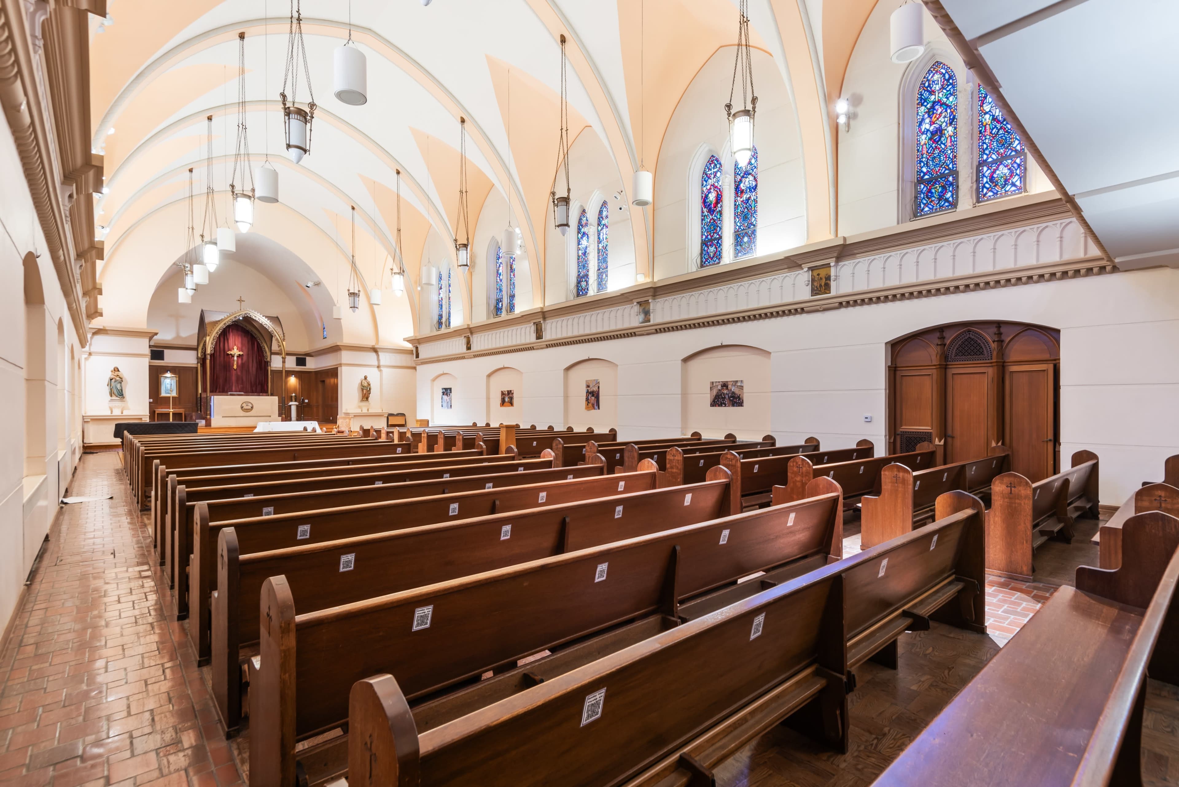 The image shows the interior of a church with wooden pews arranged in rows, stained glass windows, and a raised altar at the front.