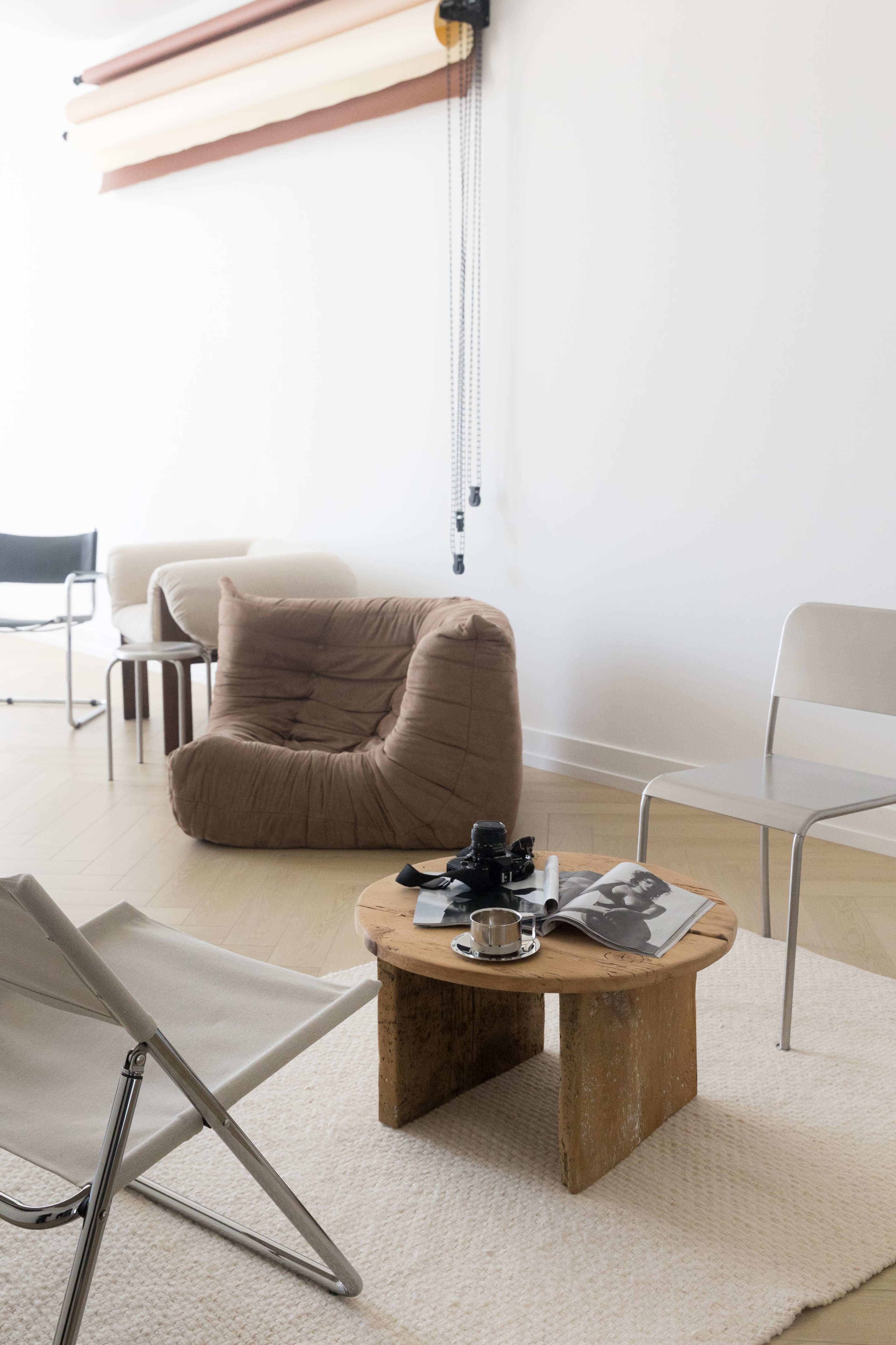 The image shows a minimalist living space featuring a wooden coffee table with a magazine and a camera, surrounded by a bean bag chair and two metal chairs on a soft rug.