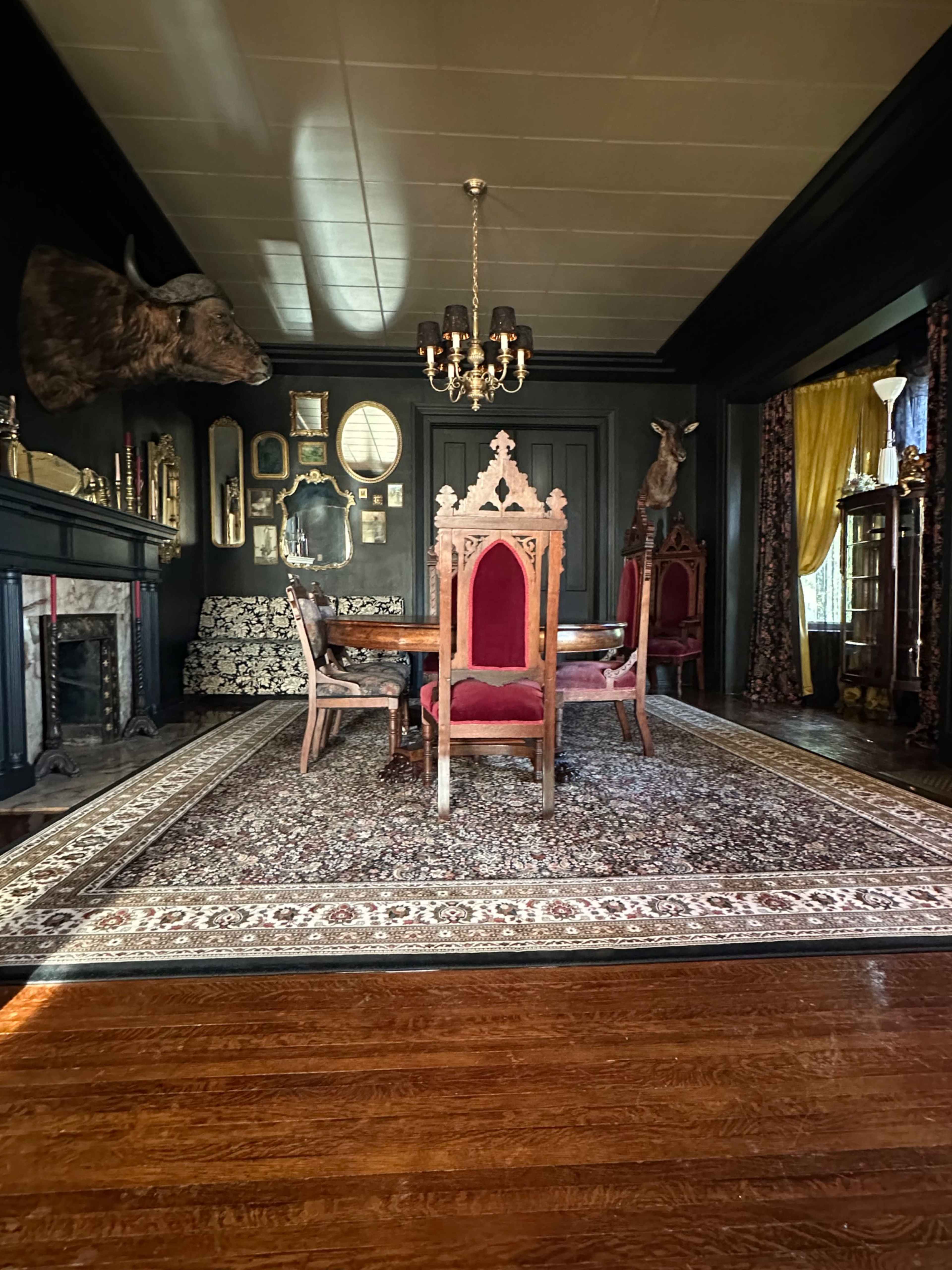 A dimly lit dining room featuring an intricately designed wooden table surrounded by high-backed chairs, a large area rug, and various decorative elements on the walls.