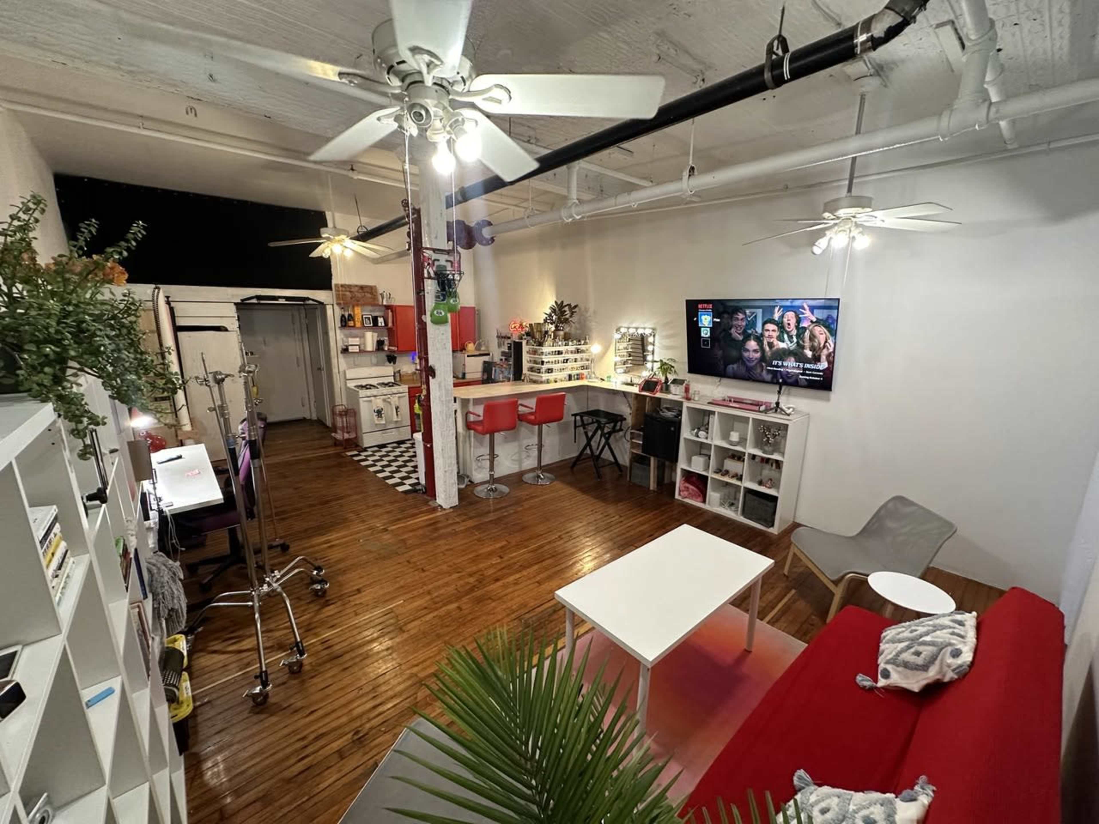 The image shows a cozy studio apartment with wooden floors, a red sofa, a white coffee table, and a kitchen area in the background, along with modern light fixtures and decorative plants.