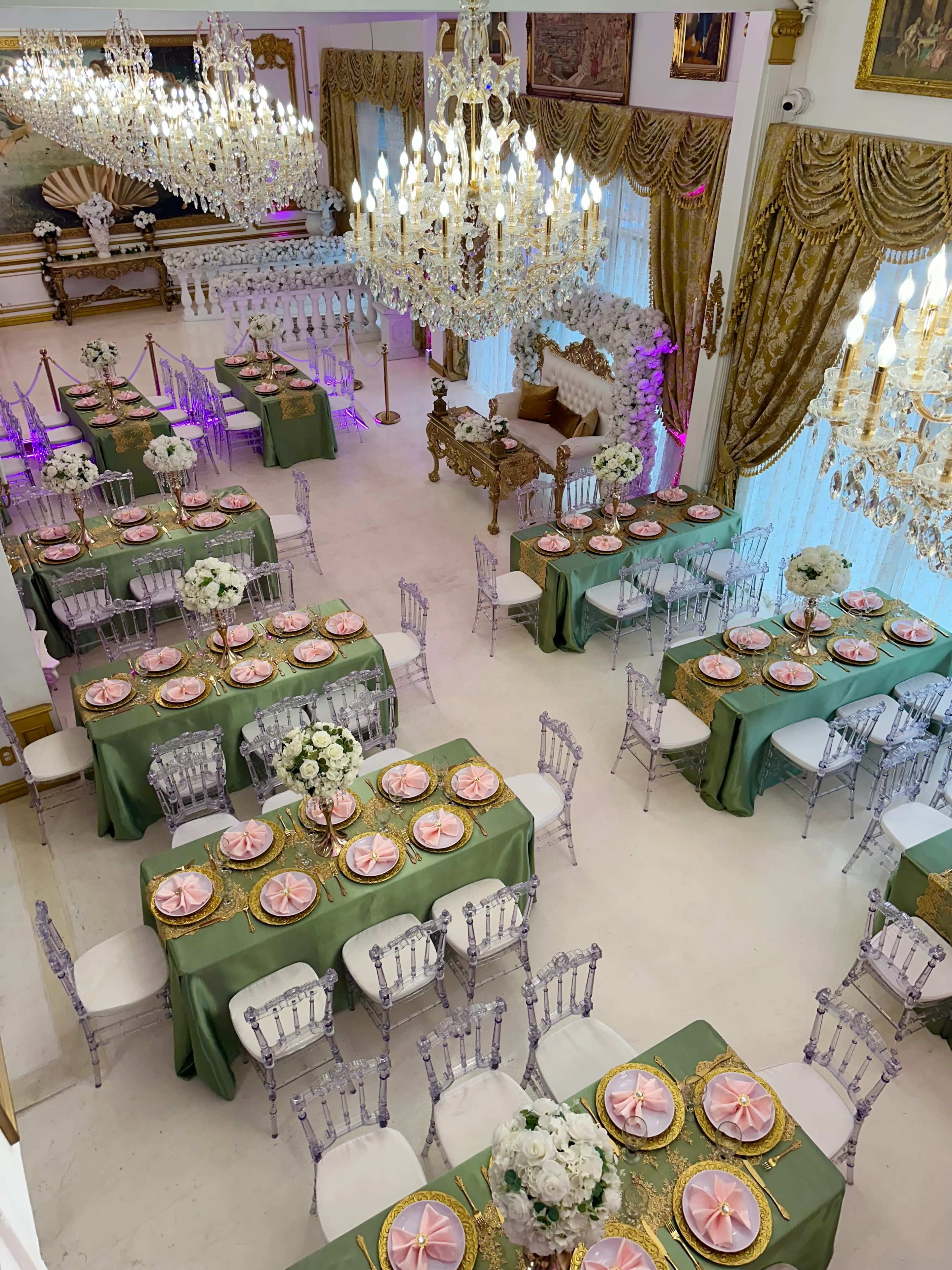 The scene shows a banquet hall set for an event, featuring elegantly arranged tables with green tablecloths, pink plates, and floral centerpieces under grand chandeliers.