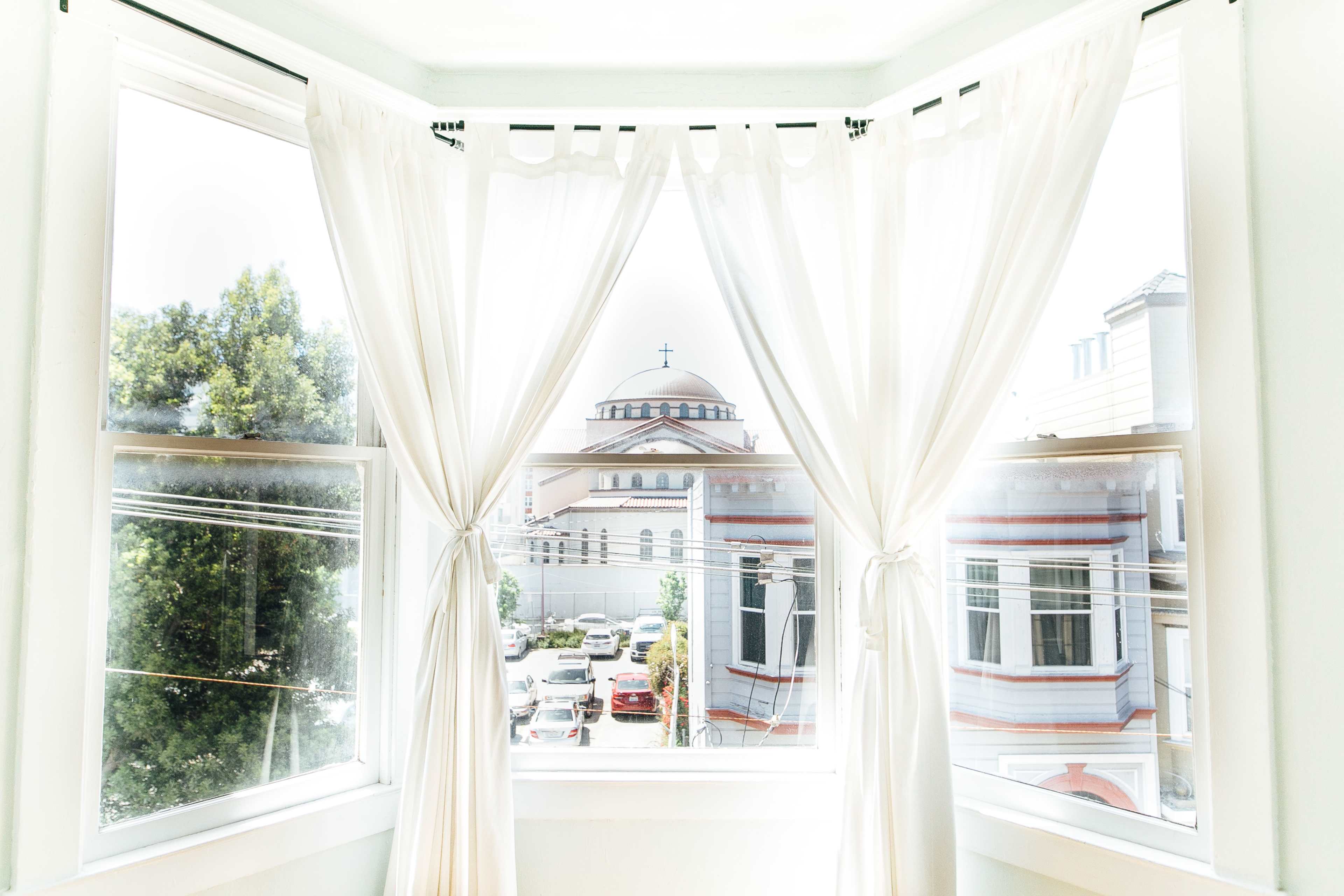 A bright window with sheer white curtains, framing a view of a building with a dome and cross in the distance, surrounded by parked cars and trees.