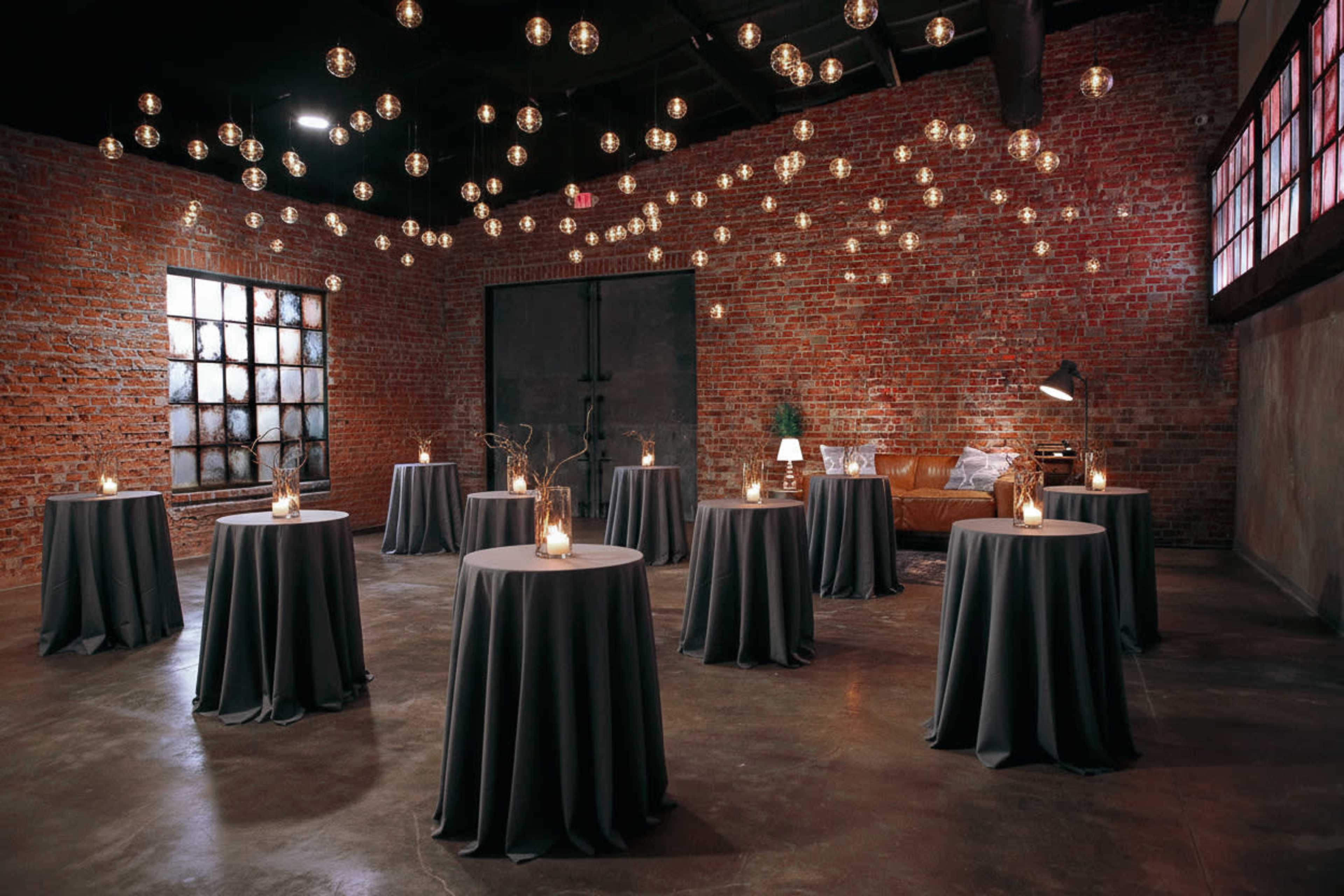 The image shows a spacious brick-walled room with multiple round tables covered in dark cloth, each adorned with candles and decorative elements, and illuminated by hanging light fixtures.