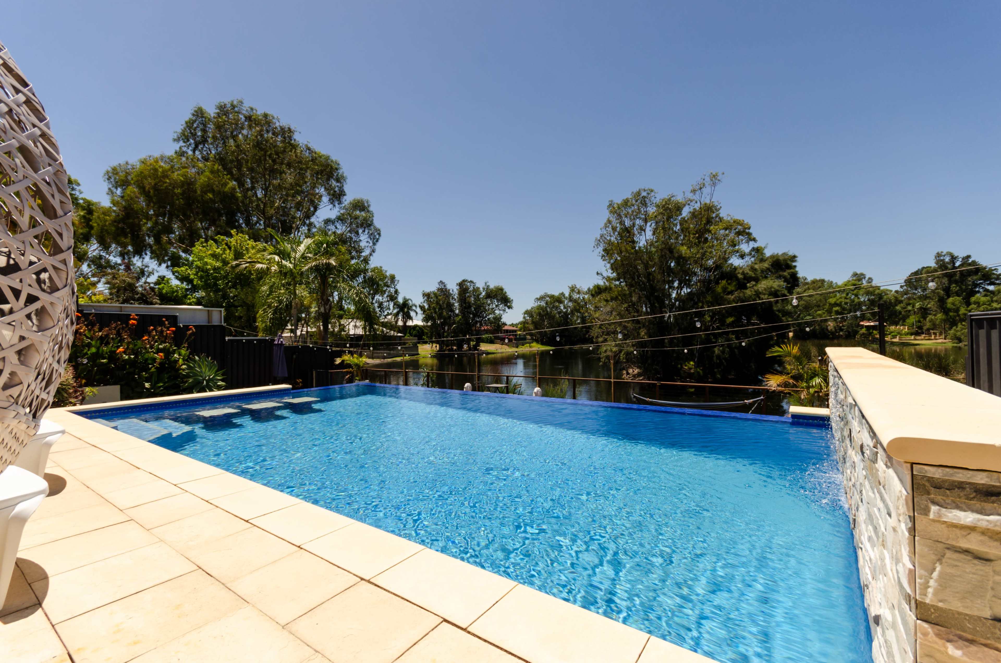 A clear swimming pool reflects the blue sky, surrounded by greenery and overlooking a calm waterway.