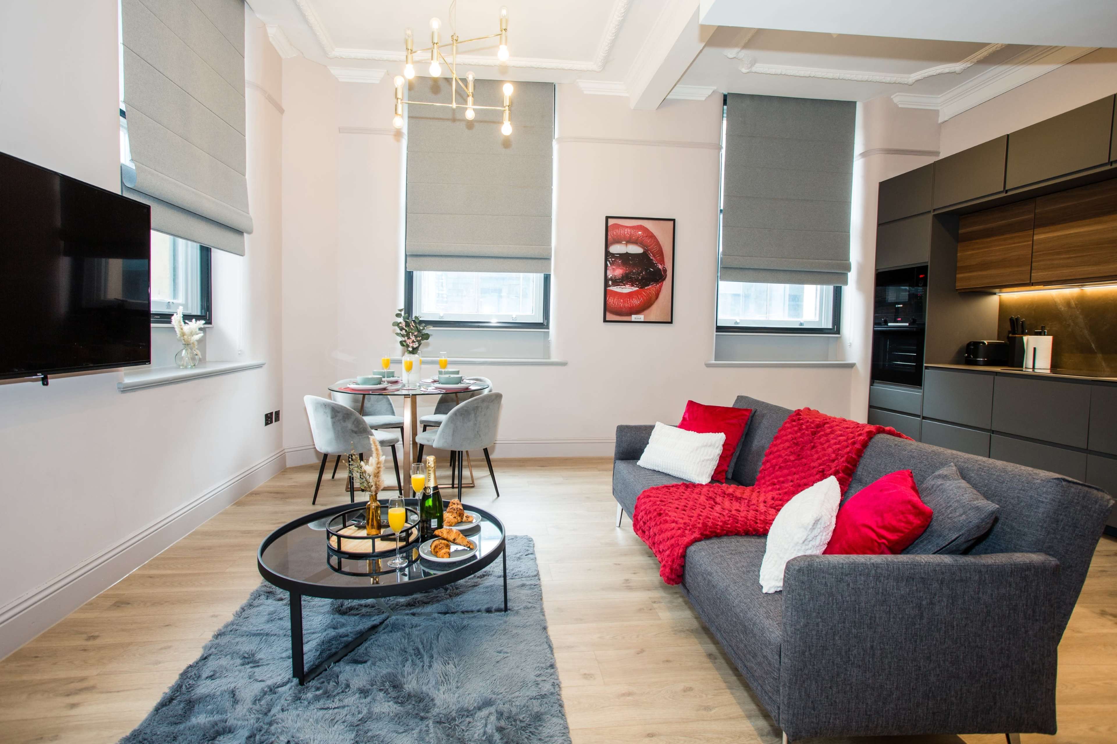 The image shows a modern living space with a gray sofa, a round dining table set for two, and an open kitchen area, all decorated with contemporary furnishings and artwork.