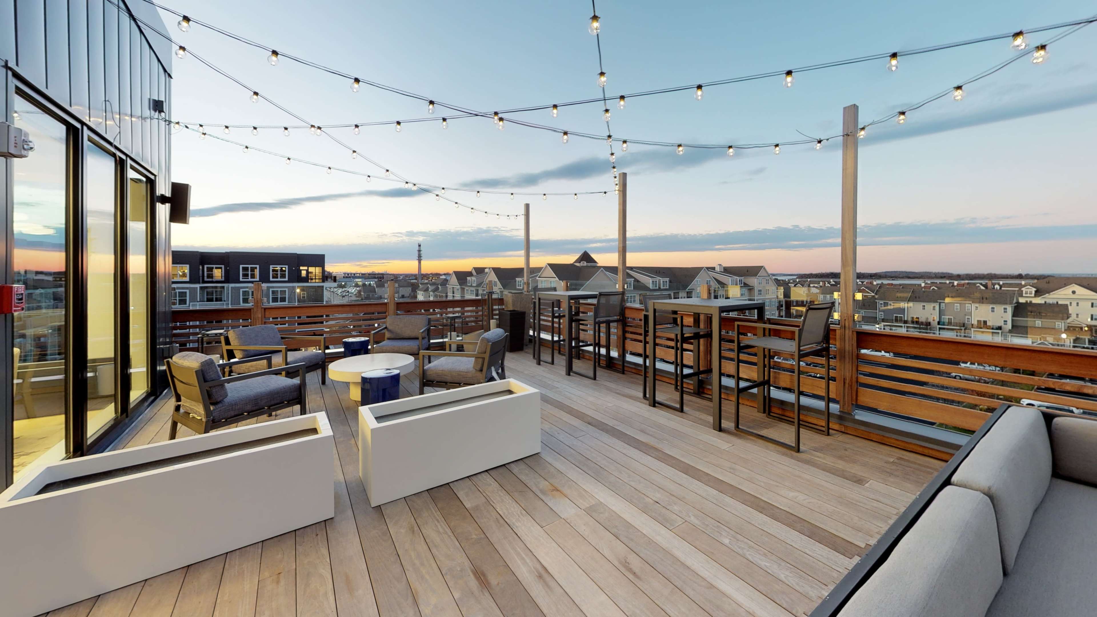 A rooftop terrace with wooden flooring, string lights overhead, and various seating arrangements, including armchairs and tables, overlooking a residential area at sunset.
