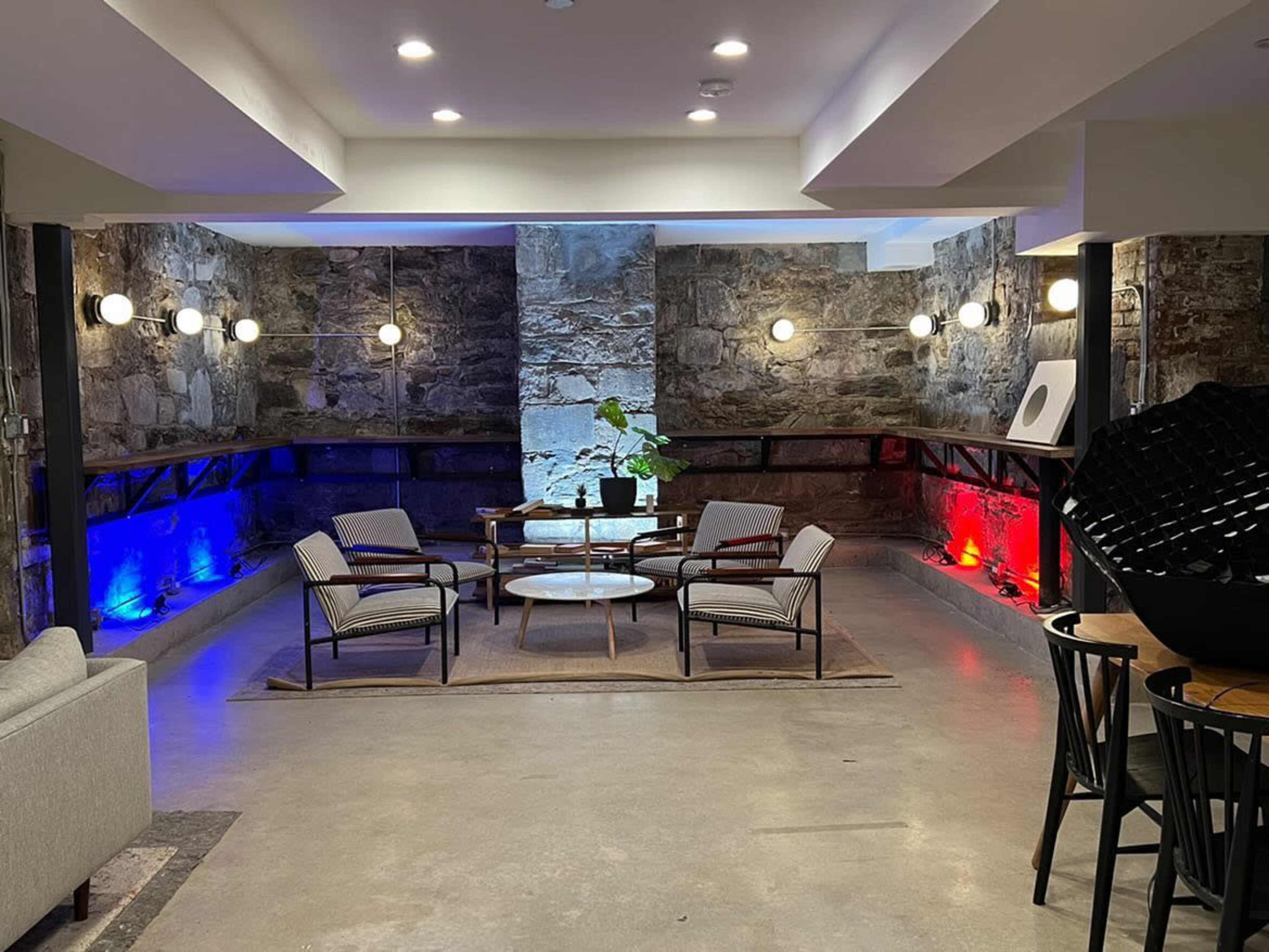 A modern seating area with four chairs and a glass table is illuminated by wall-mounted lights and colorful accents against a stone wall backdrop.