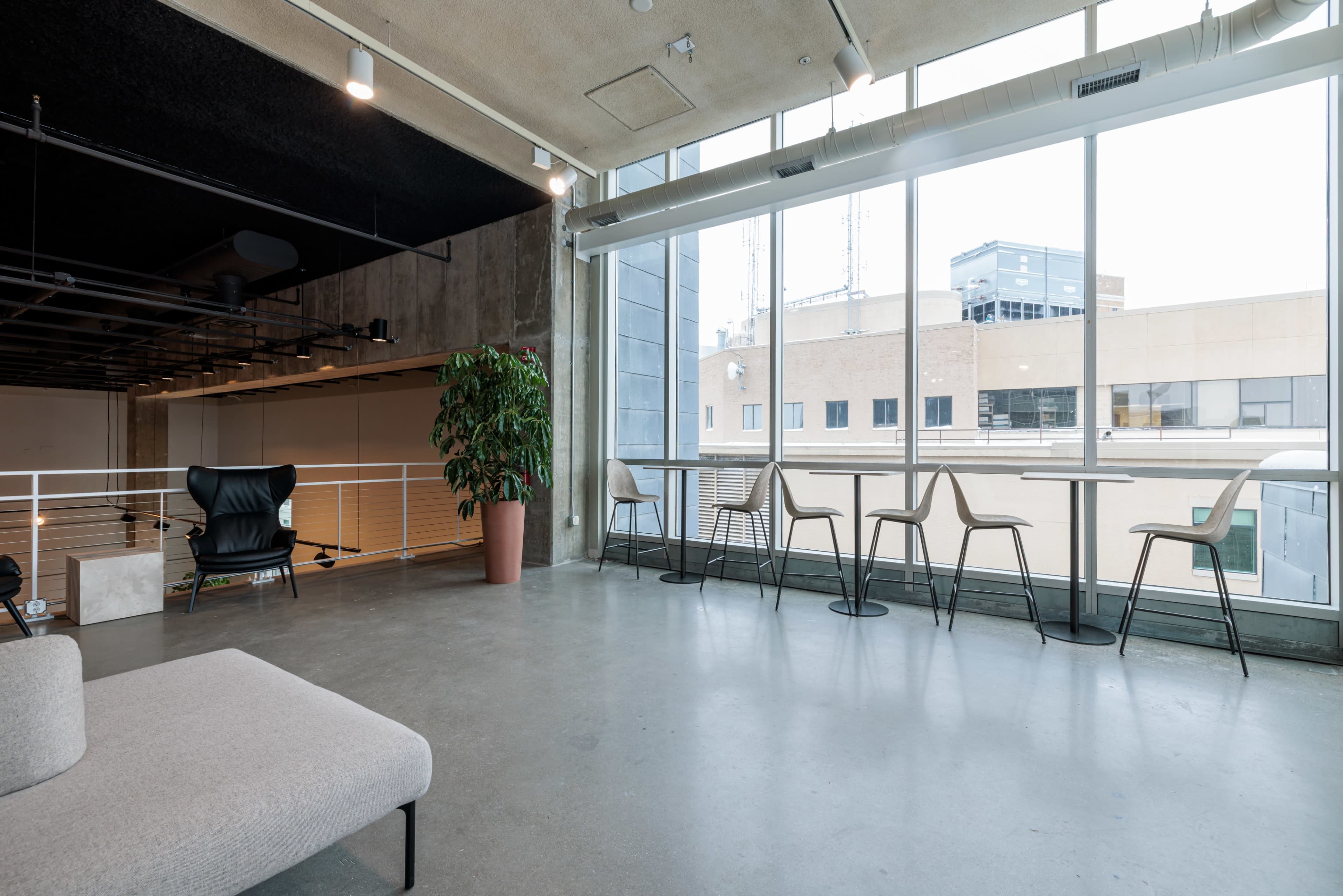 The image shows a modern interior space with a gray sofa, several chairs, and large windows overlooking a cityscape.