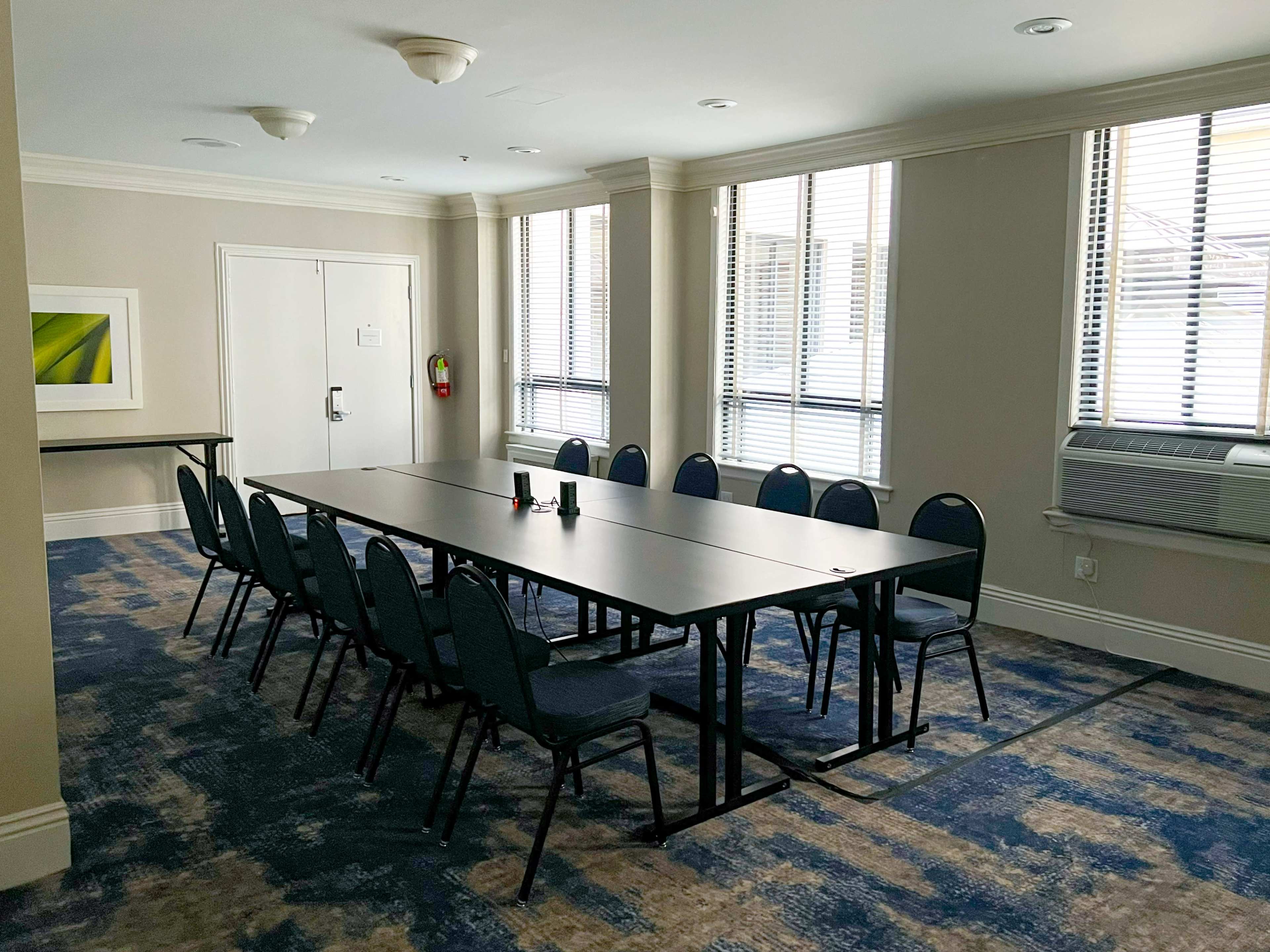 The image shows a conference room with a long table and several chairs arranged around it, featuring windows that let in natural light.