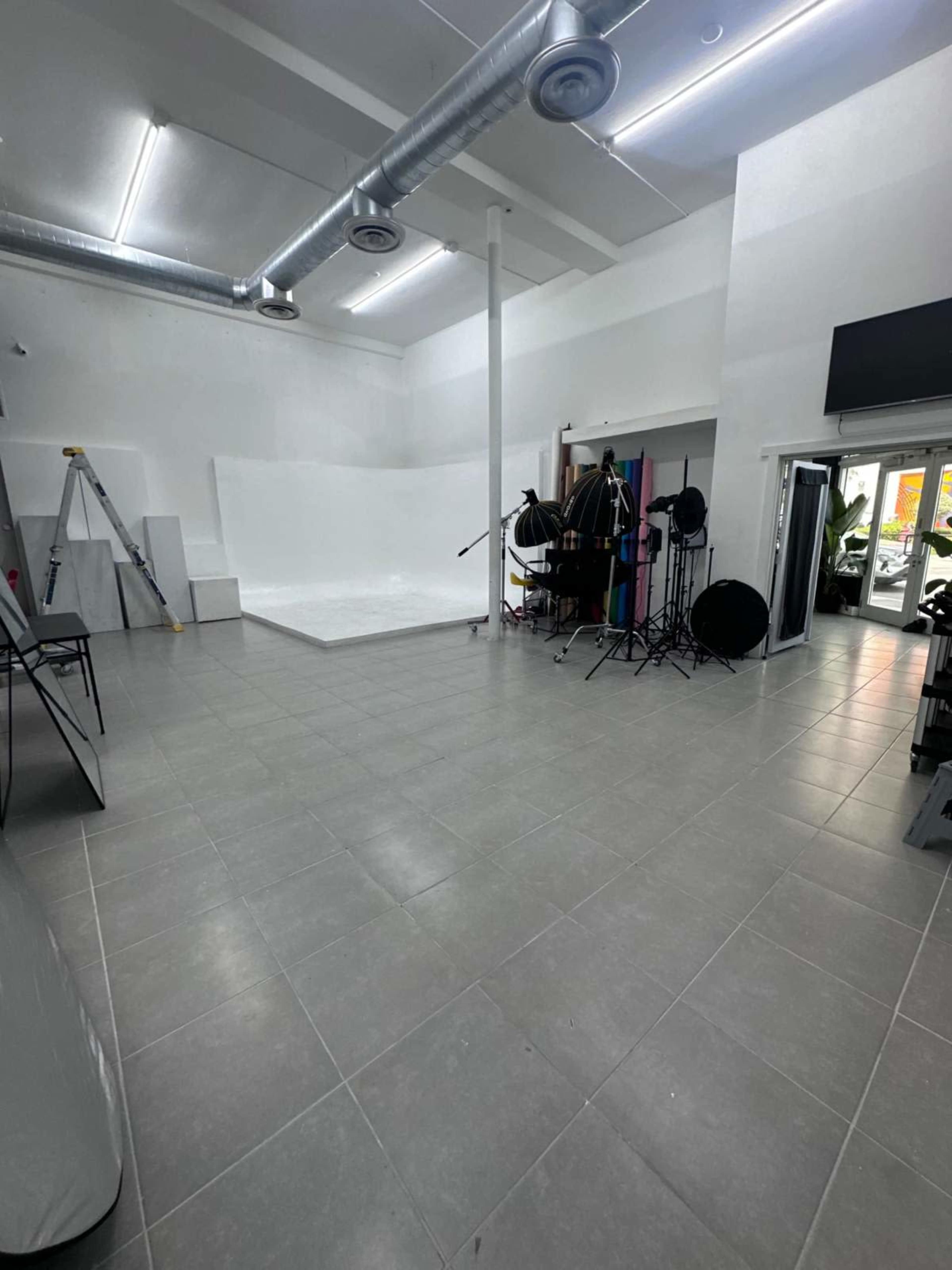 The image shows a spacious photo studio with a white backdrop, lighting equipment, and various props arranged around the room.