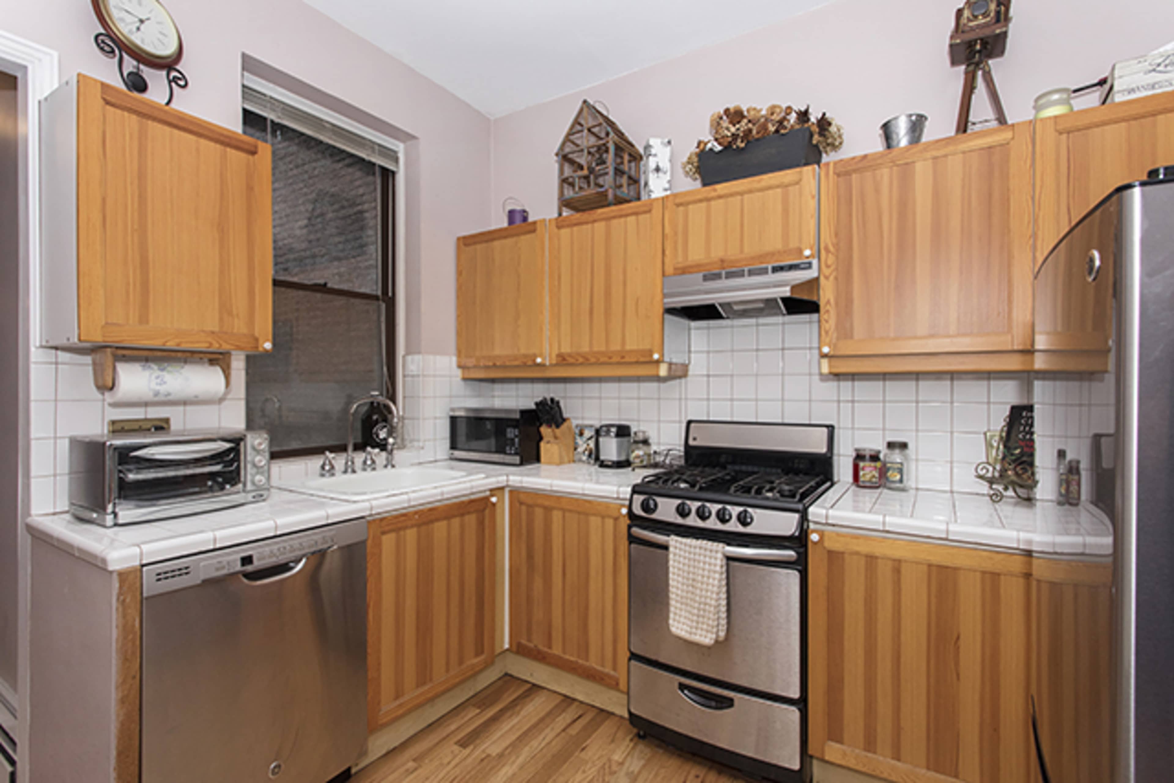 The image shows a kitchen with wooden cabinets, a stainless steel stove, and white tiled countertops.