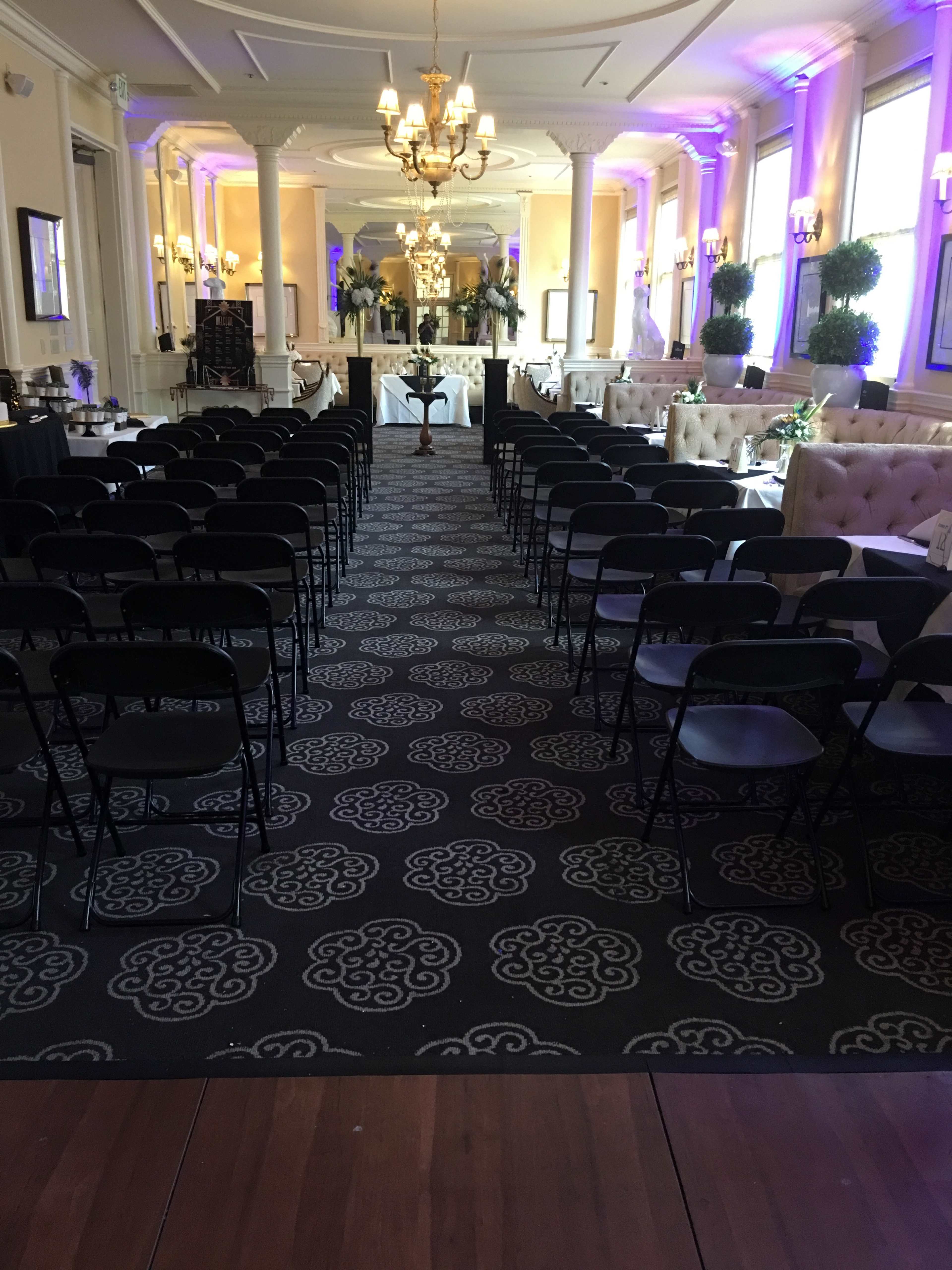 A formal indoor venue set up for an event, featuring rows of black chairs facing a decorated stage area and elegant lighting.