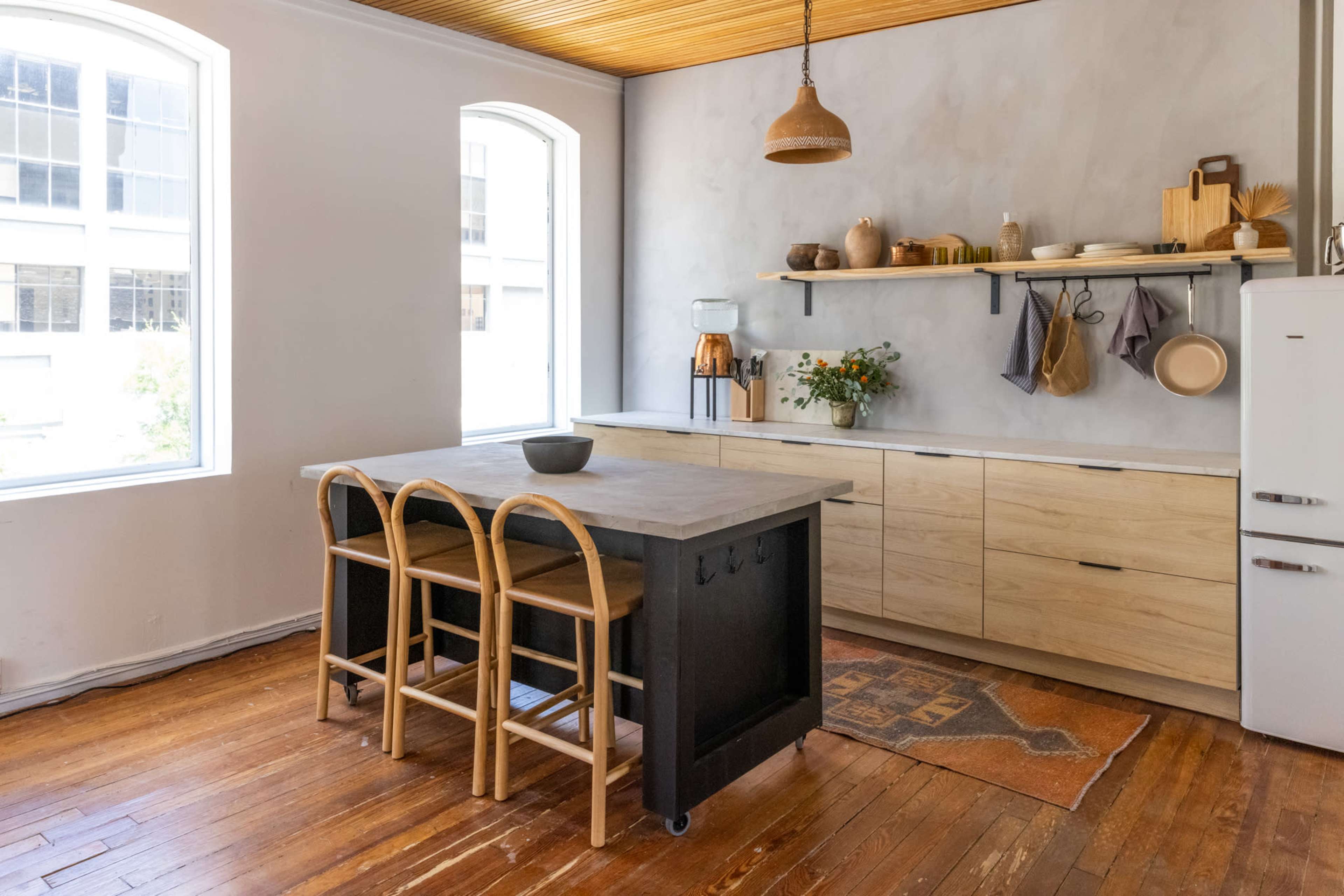 The kitchen features a wooden island with three rattan stools, modern cabinetry, and large windows that let in natural light.