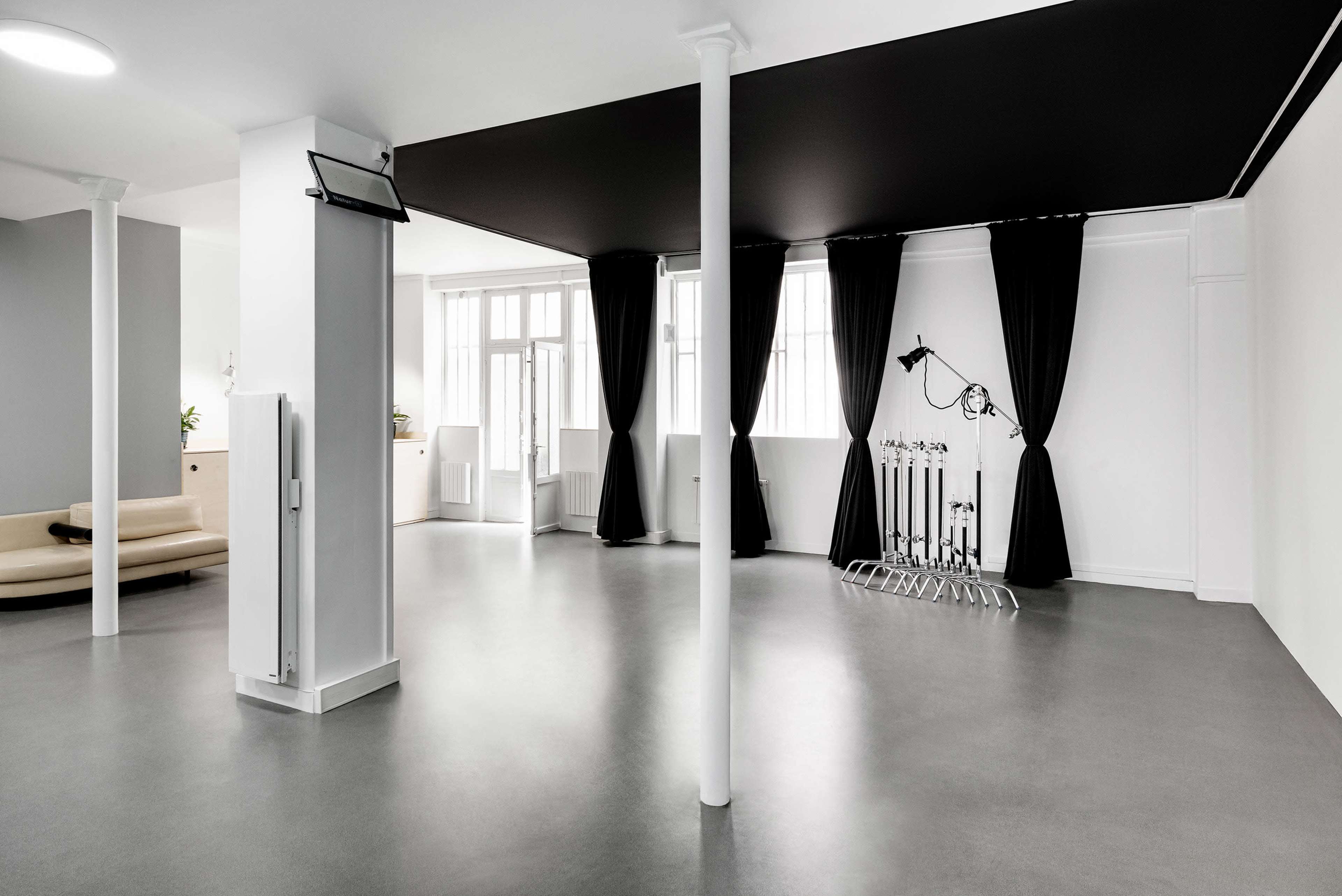 A spacious, minimalistic studio features white walls, a black ceiling, large windows, and a curtained area with equipment stands.