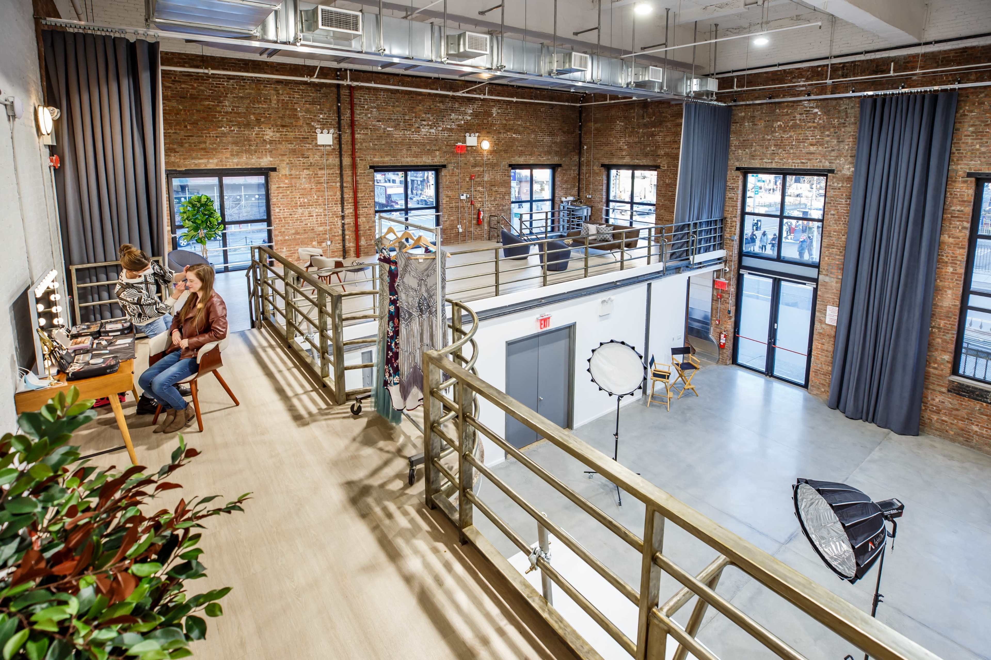 A spacious, industrial-style studio with brick walls, a second-floor walkway, and a makeup station where two women are seated.