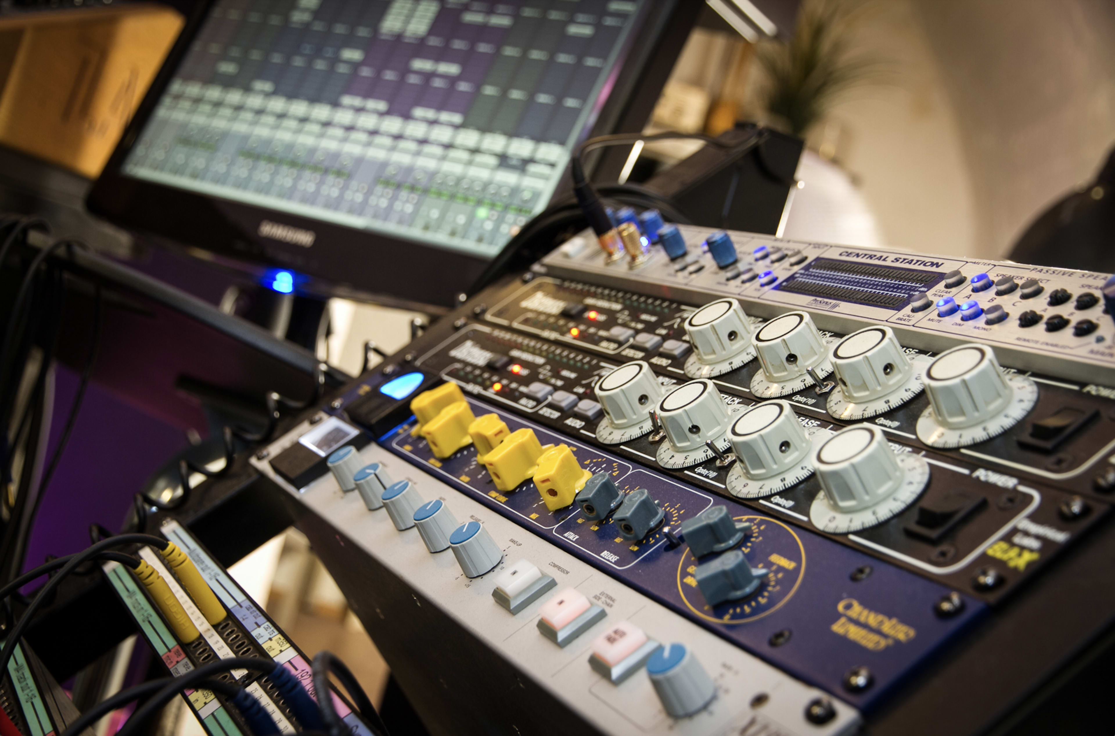 The image shows a close-up of audio mixing equipment featuring knobs and sliders alongside a computer screen displaying digital audio tracks.