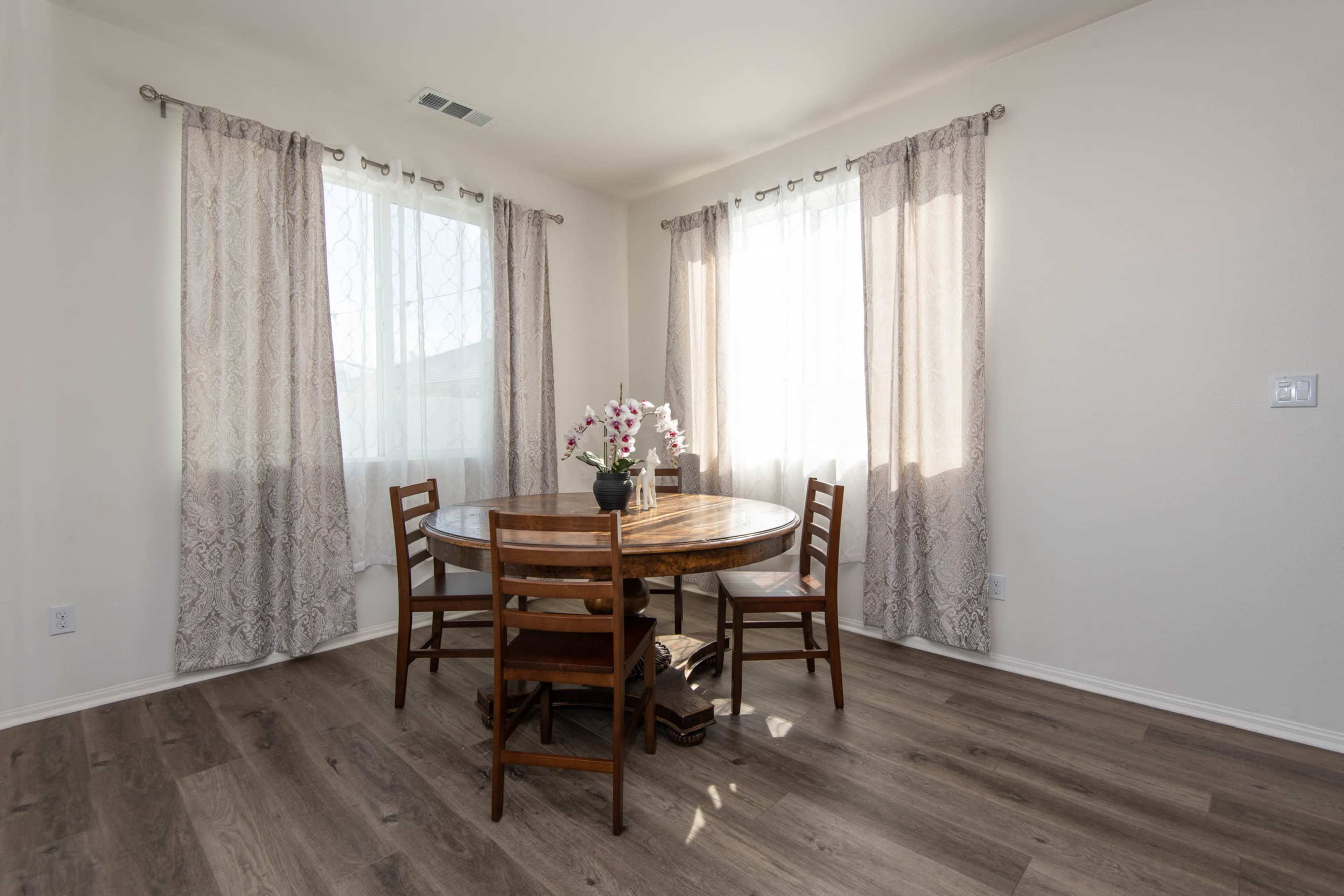 A round wooden table with chairs is positioned in a well-lit dining area, adorned with a vase of flowers and white curtains.