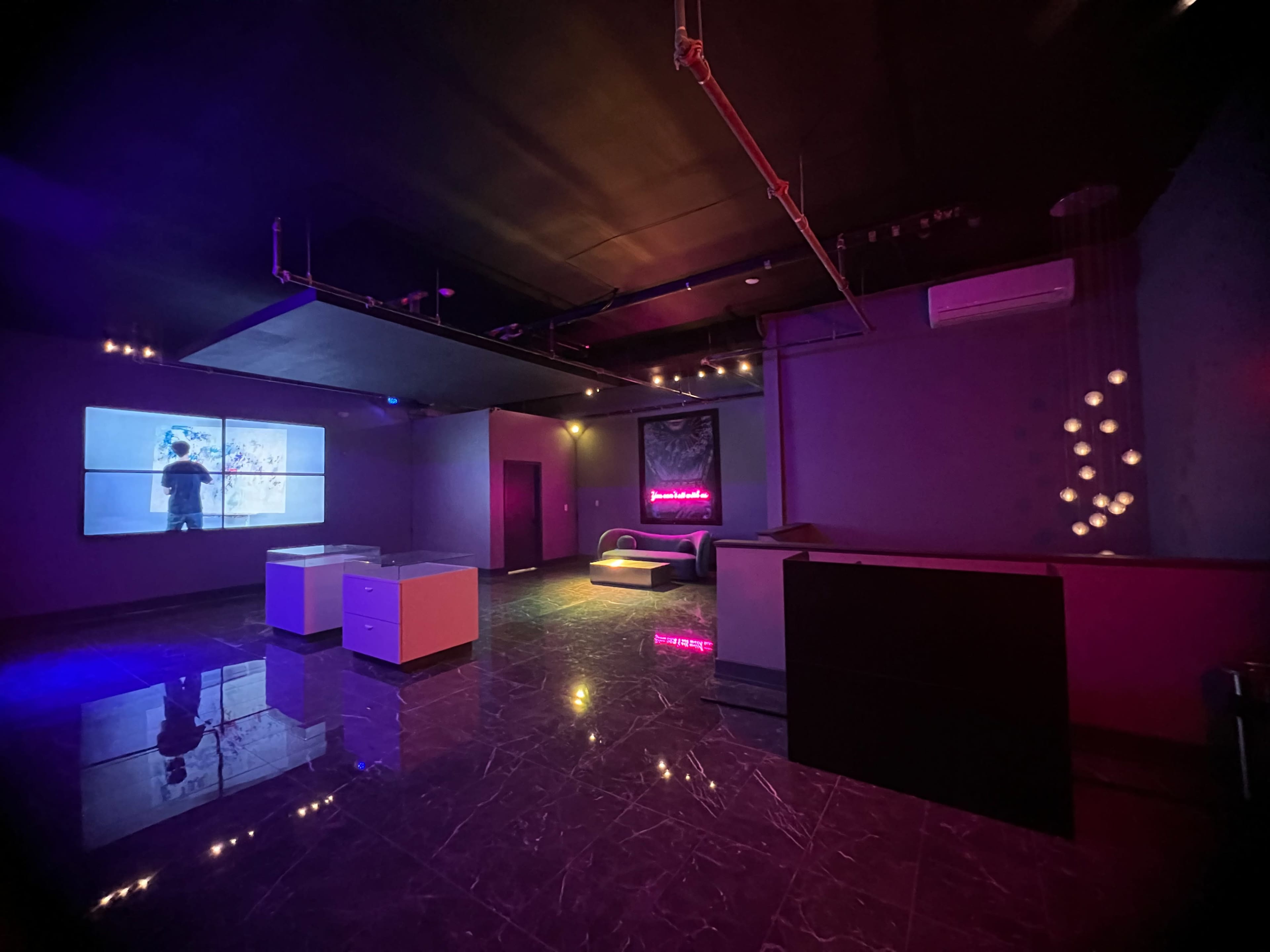 The interior of a dimly lit room features a large screen displaying a figure, modern furniture, and colorful lighting effects.