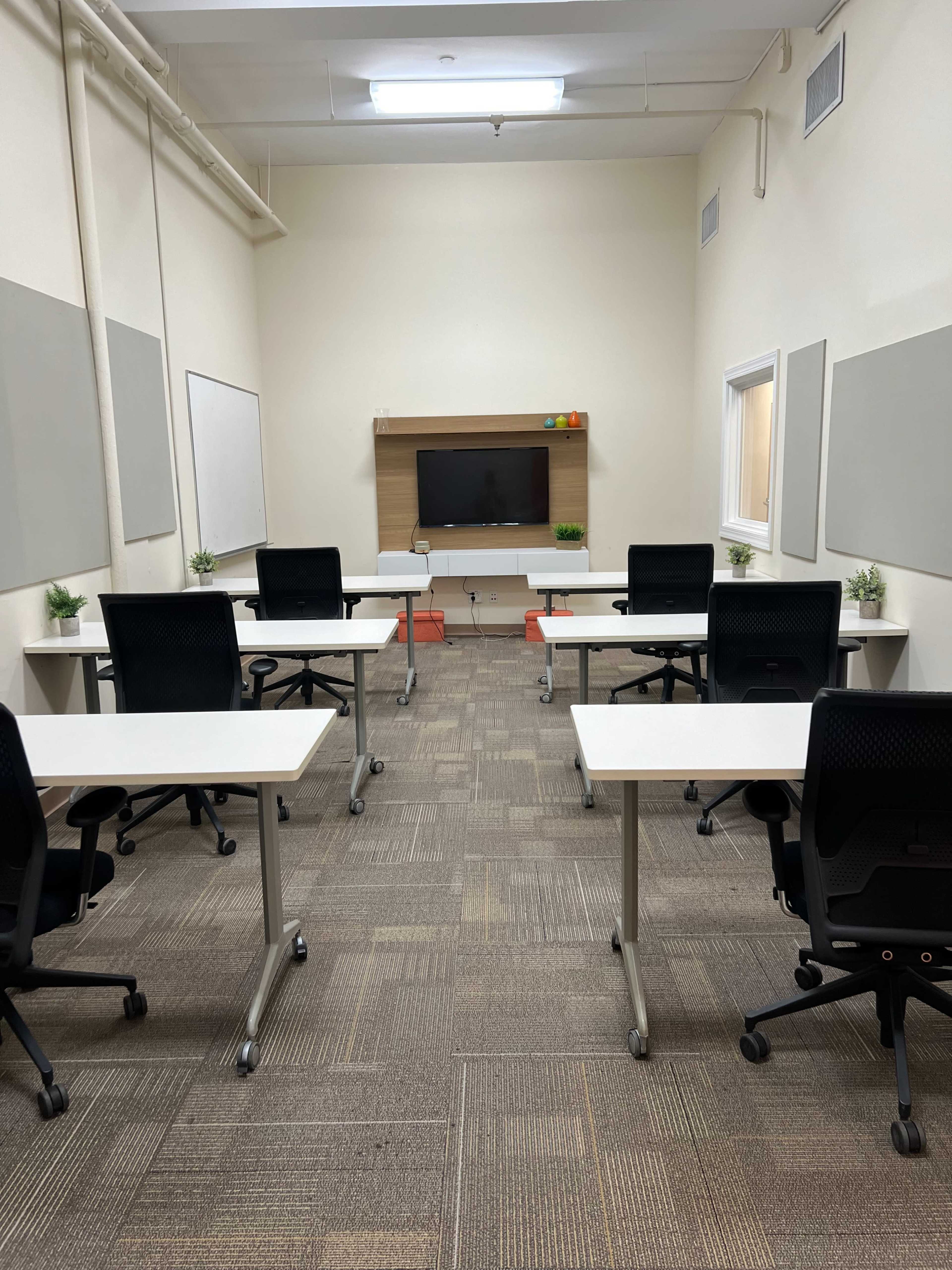 A spacious classroom features several desks and chairs arranged around a central television display and two plants on the tables.