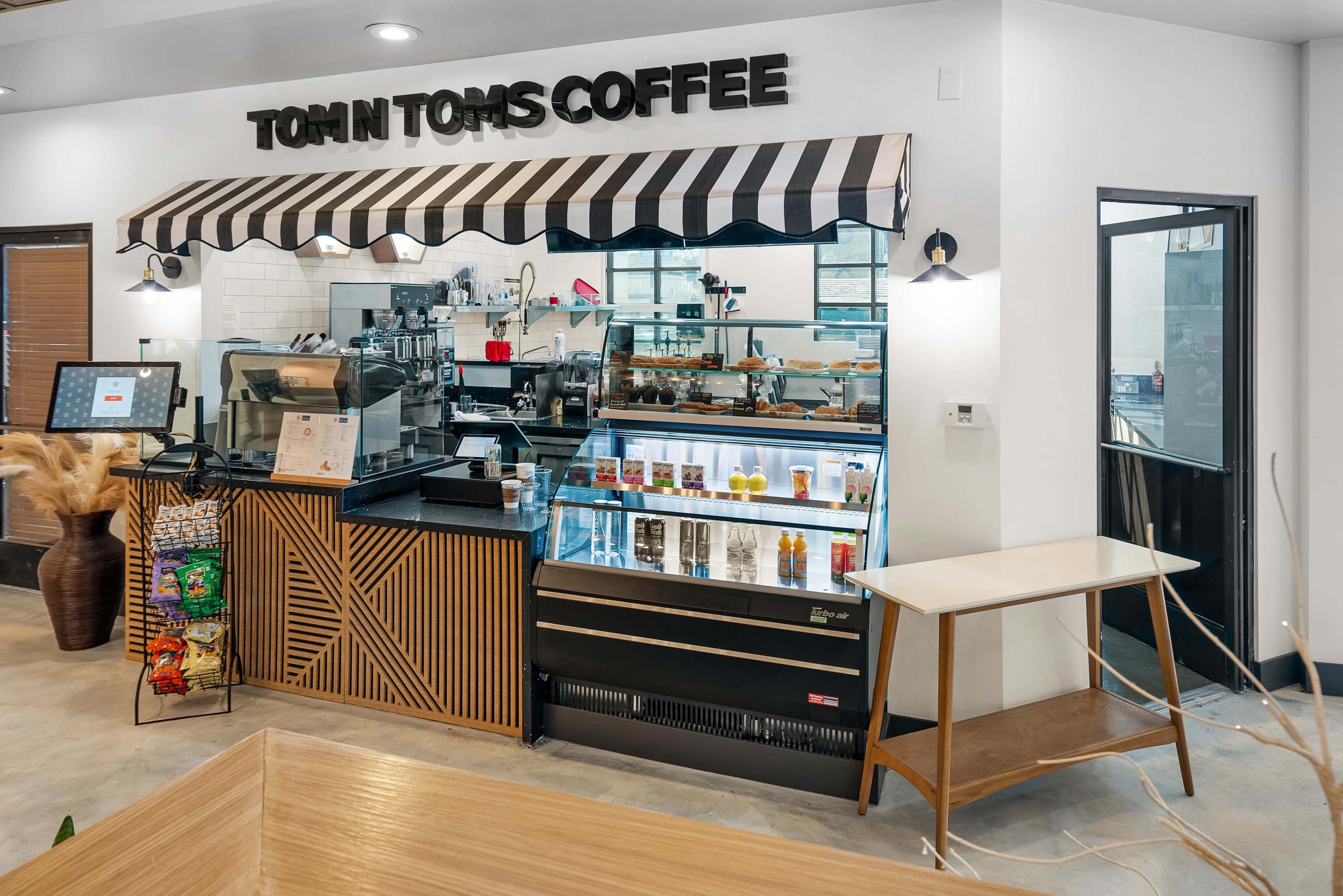 A Tom N Toms Coffee shop counter with a striped awning, a display case filled with beverages, and a small table beside it.