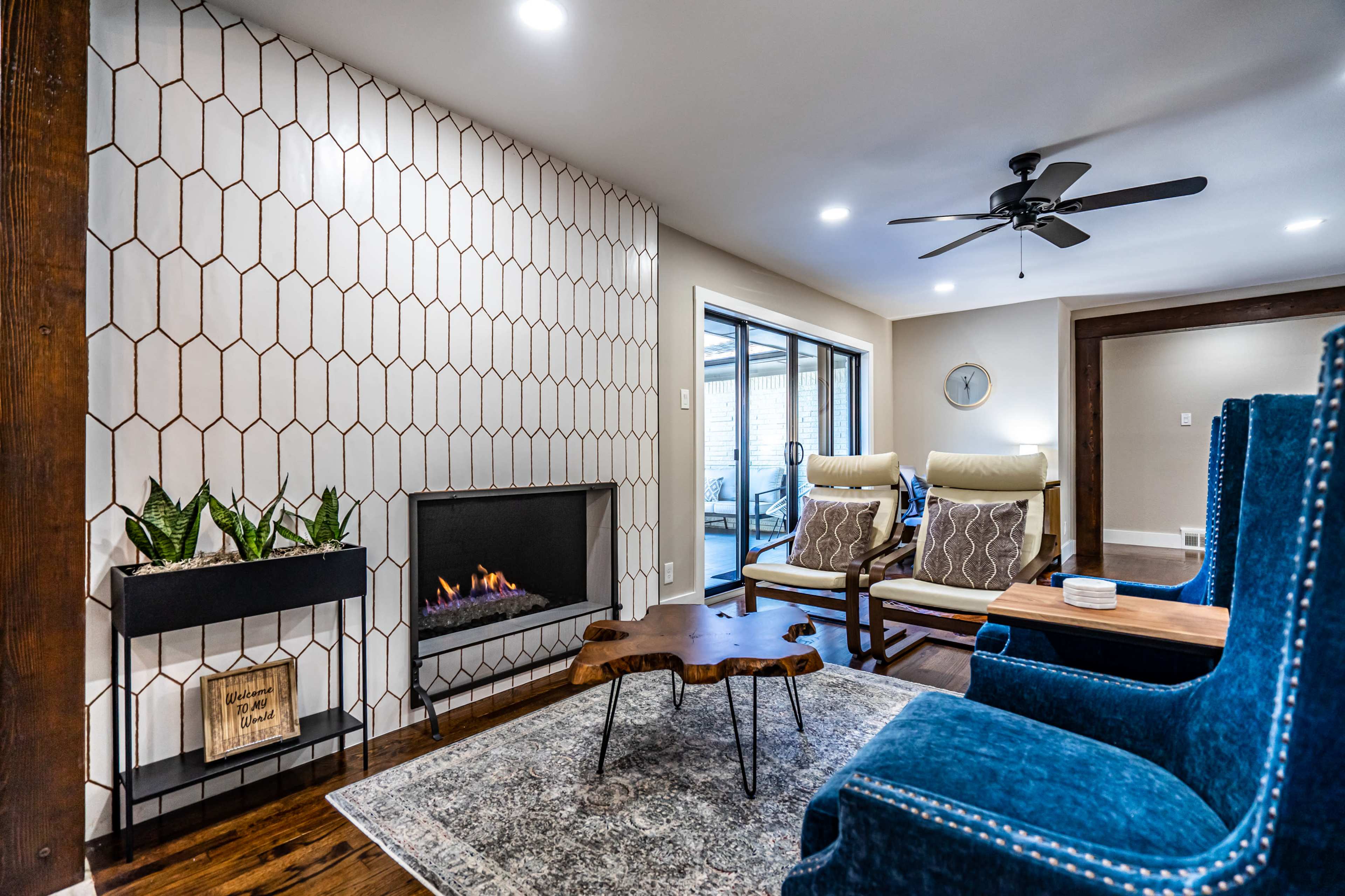 A living room with a modern fireplace, two upholstered chairs, a coffee table, and a patterned wall, with large windows providing natural light.
