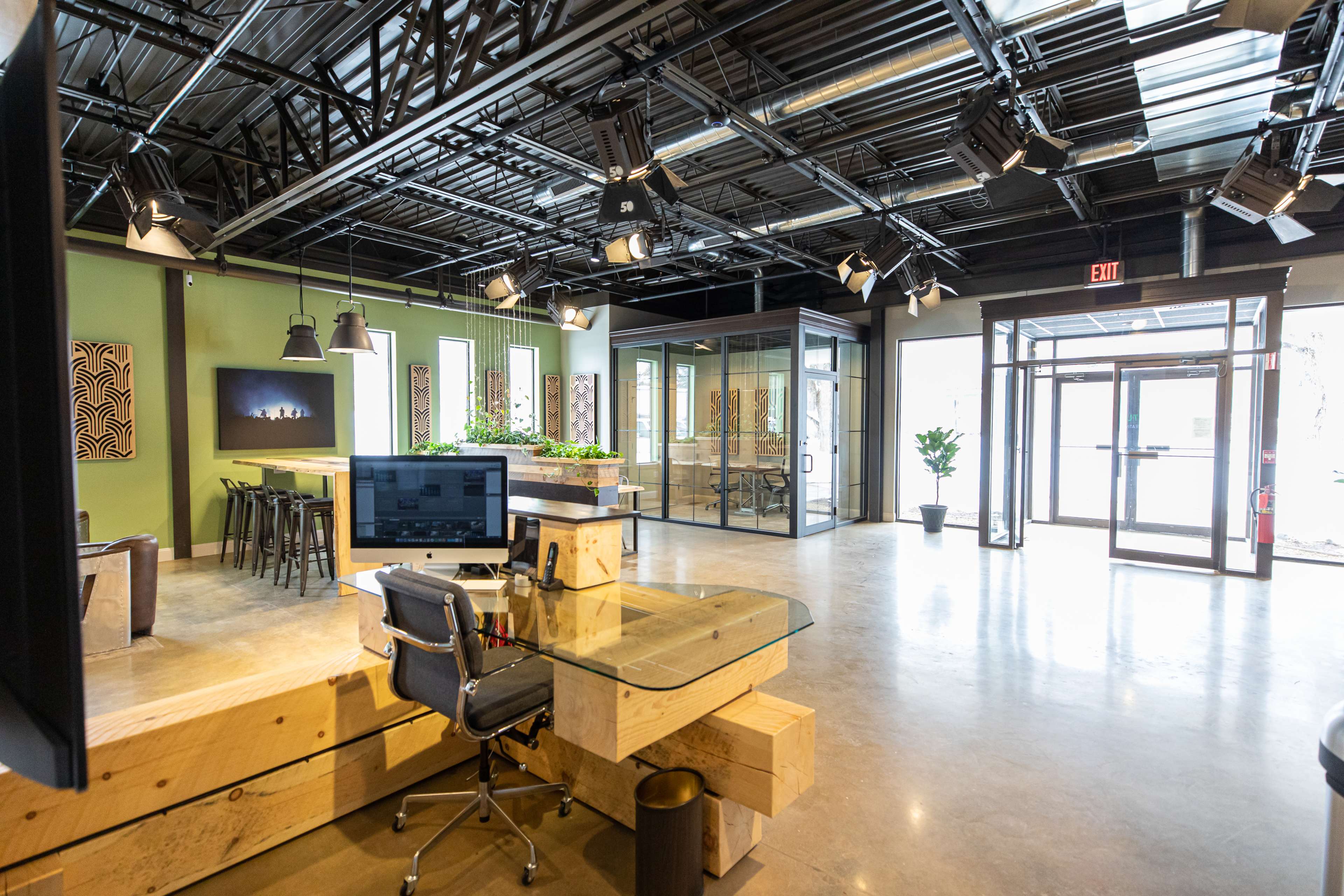 The image shows a modern office space featuring a desk with a computer, a seating area with bar stools, and glass-walled meeting rooms.