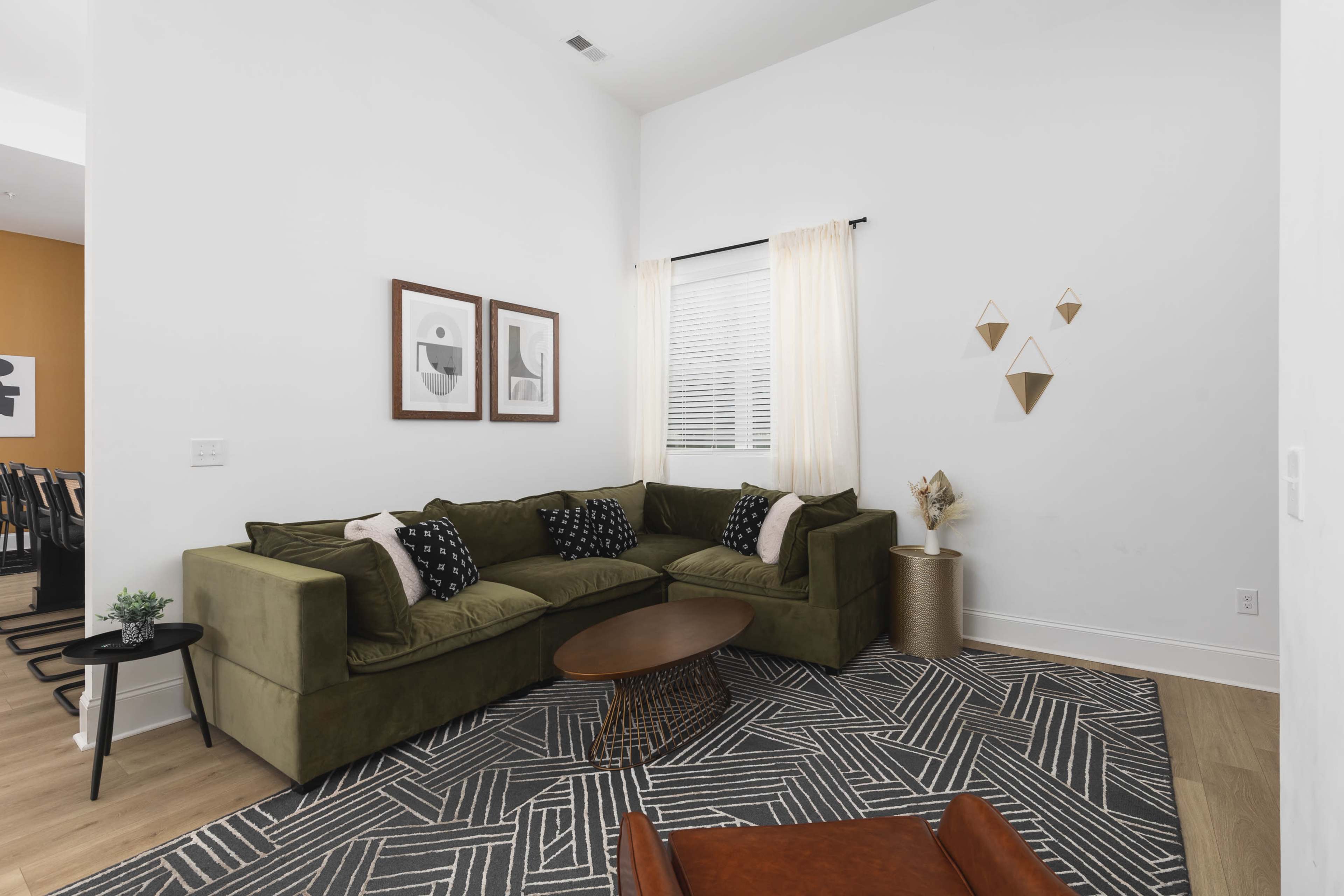 A green sectional sofa is arranged in a bright living room featuring a round coffee table, two framed artworks on the wall, and decorative items on a shelf.