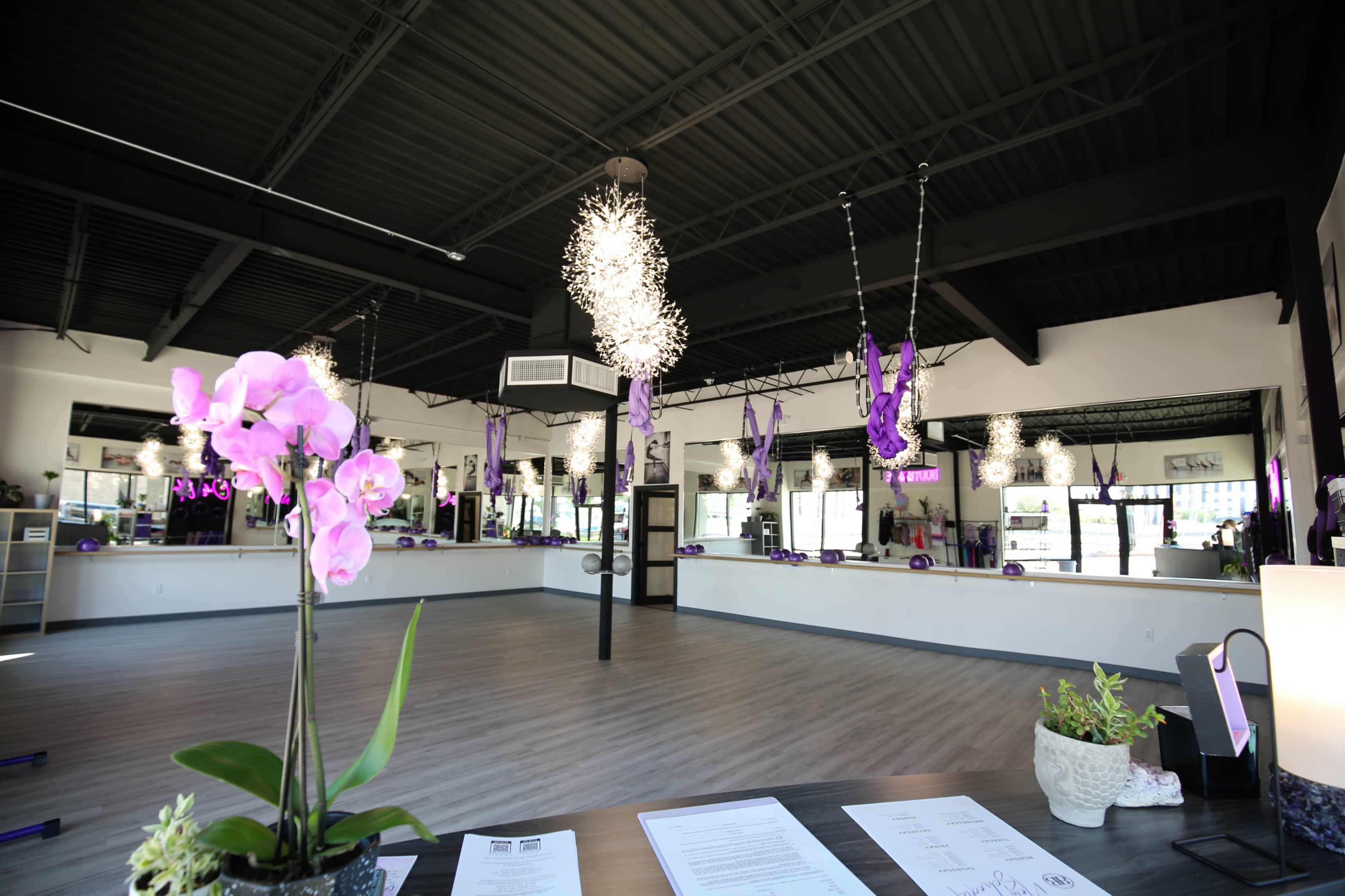The image shows a spacious interior of a fitness studio featuring purple decorations, hanging light fixtures, and a front desk with a flower arrangement.