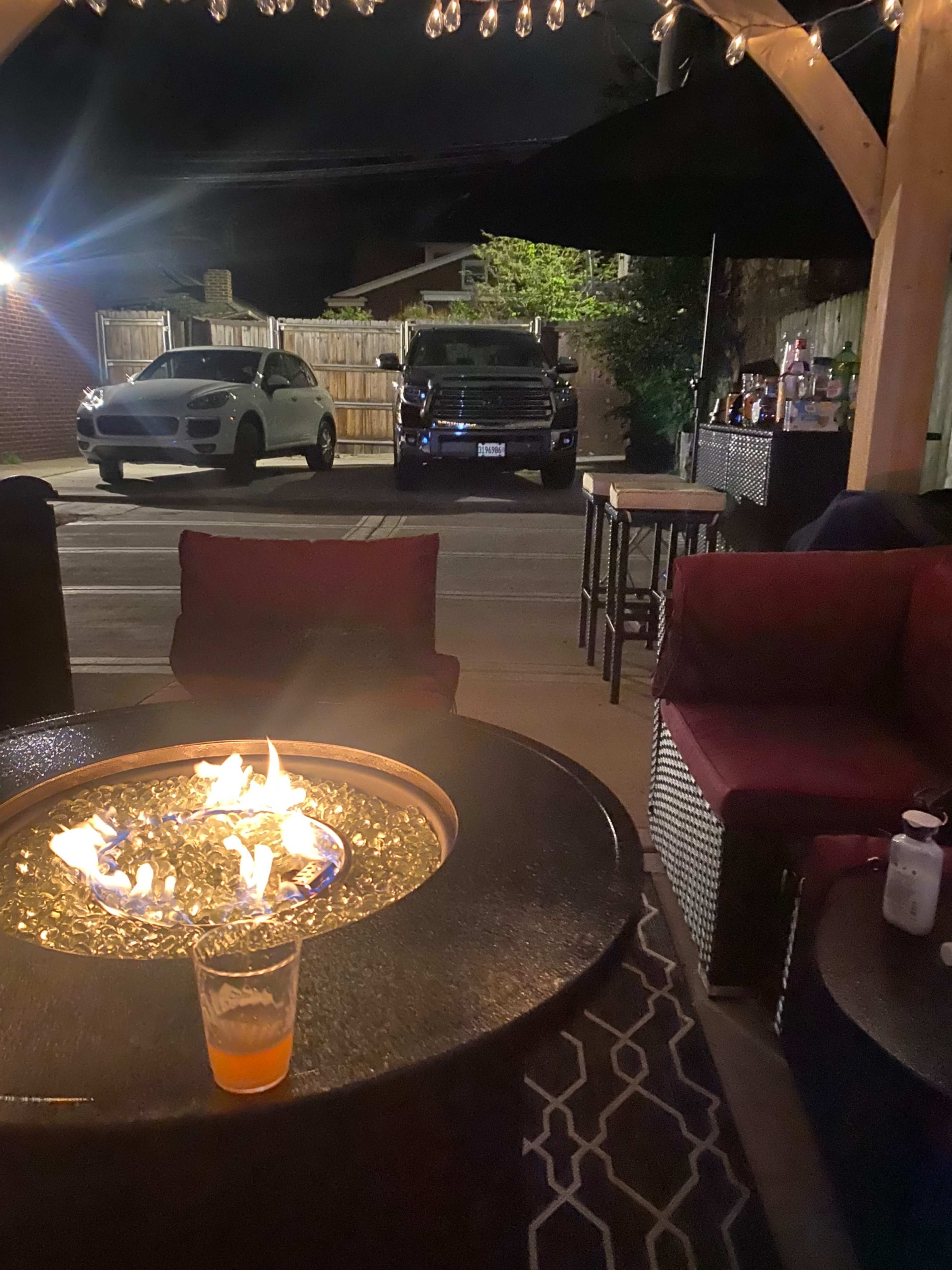 The scene shows a dimly lit outdoor area with a fire pit in the foreground, surrounded by seating, and two parked vehicles visible in the background.