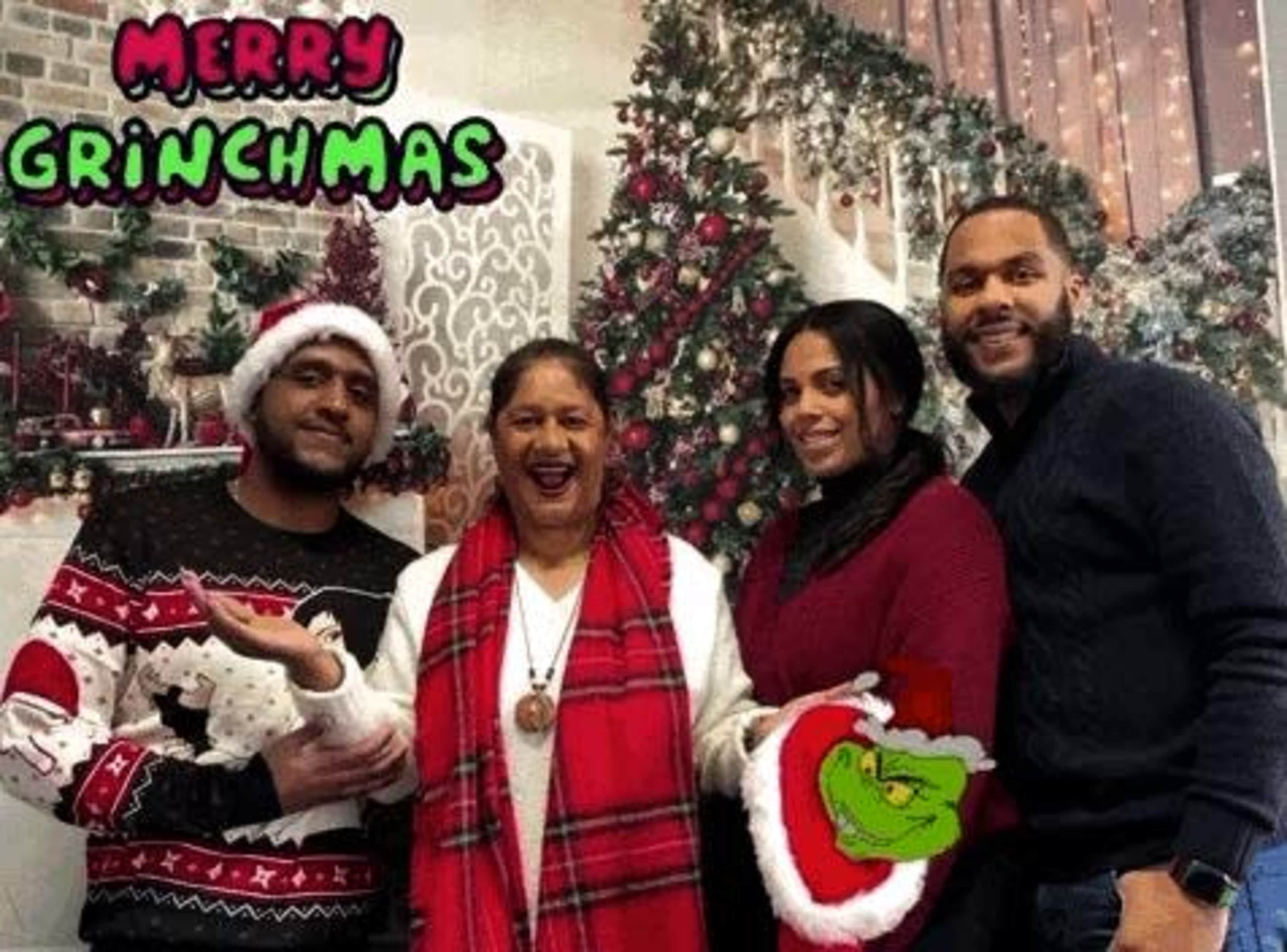 A group of four people stands together in front of a festive holiday backdrop featuring a decorated Christmas tree and a "Merry Grinchmas" graphic.