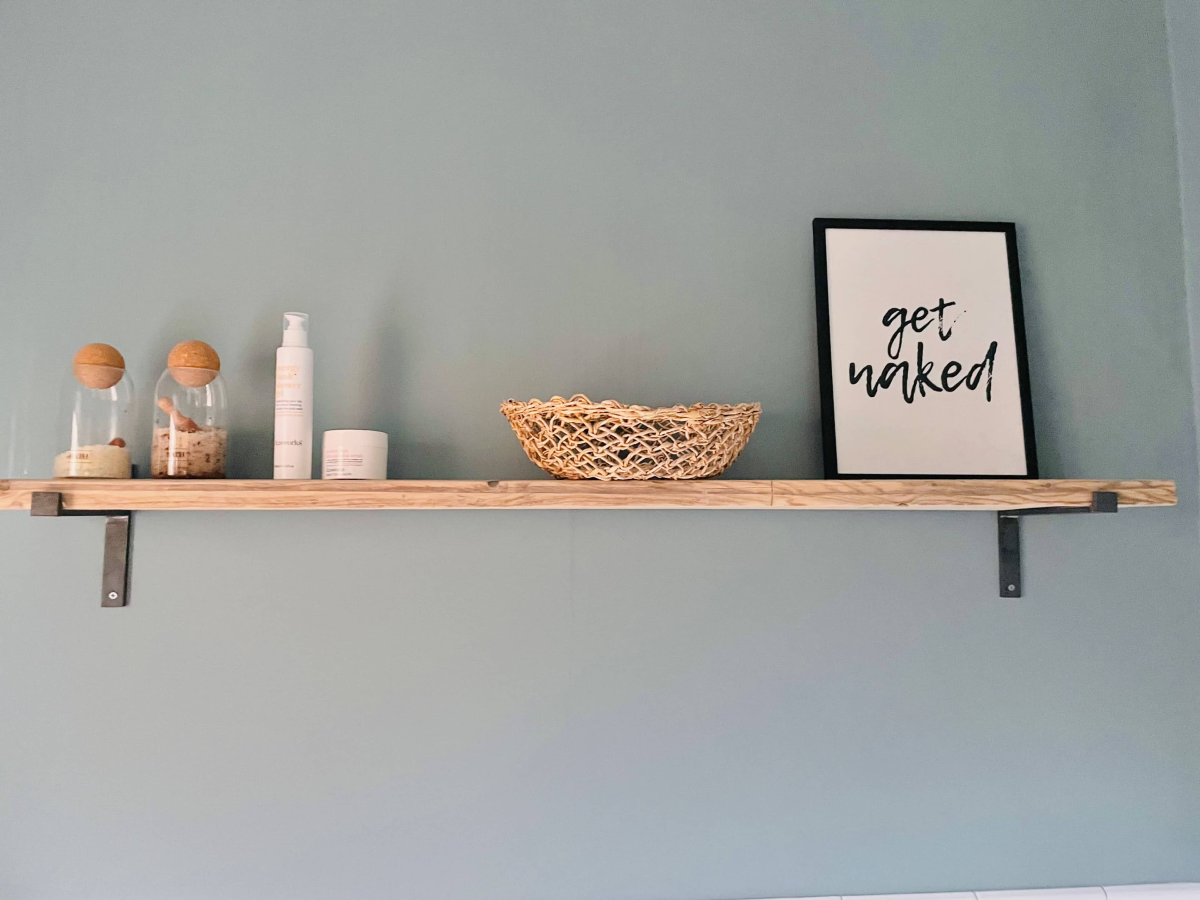 A wooden shelf mounted on a wall displays two glass containers, a lotion bottle, a woven basket, and a framed print that reads "get naked."