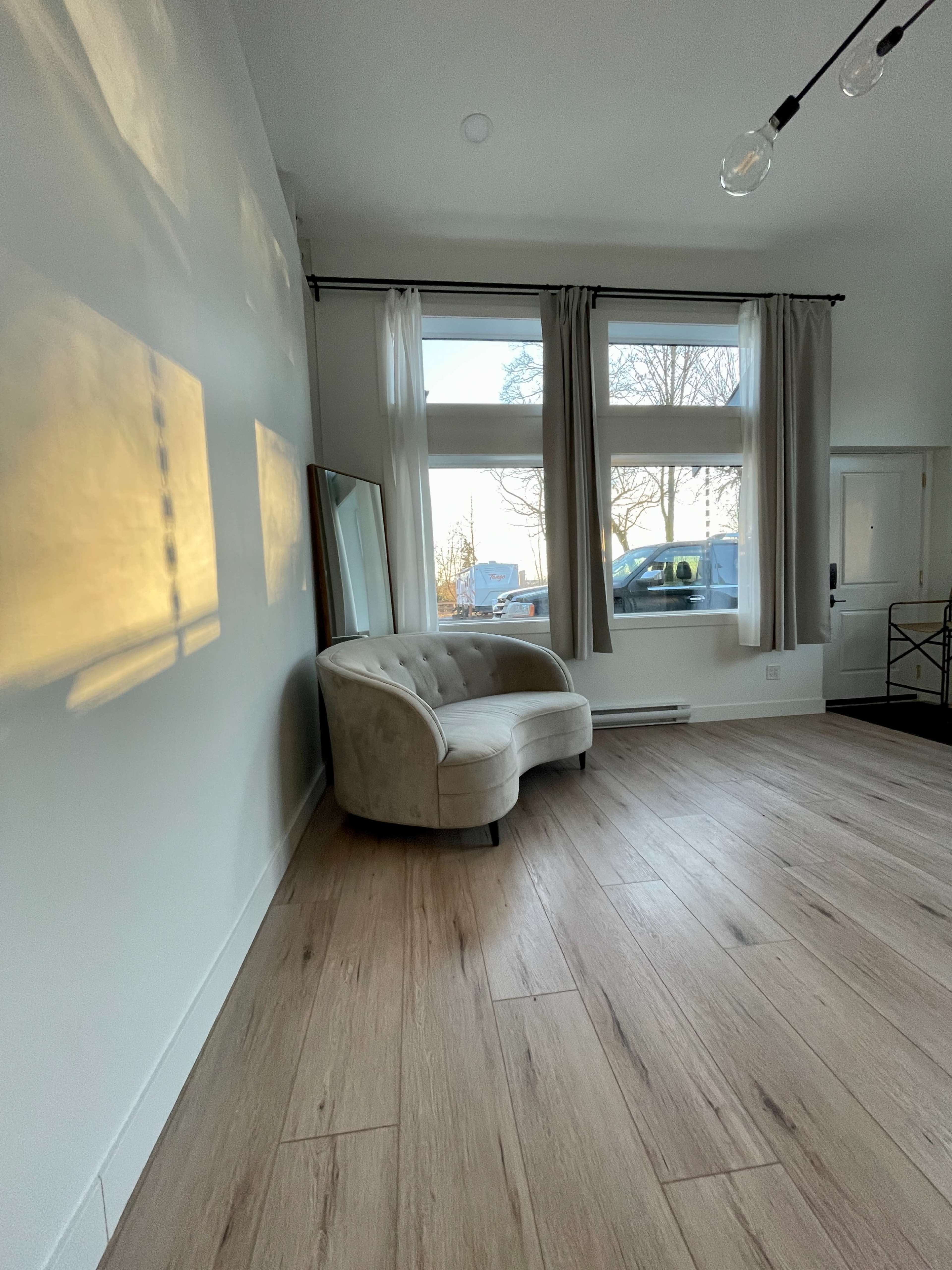 A light-colored, upholstered couch is positioned against a wall in a room with large windows and wooden flooring.