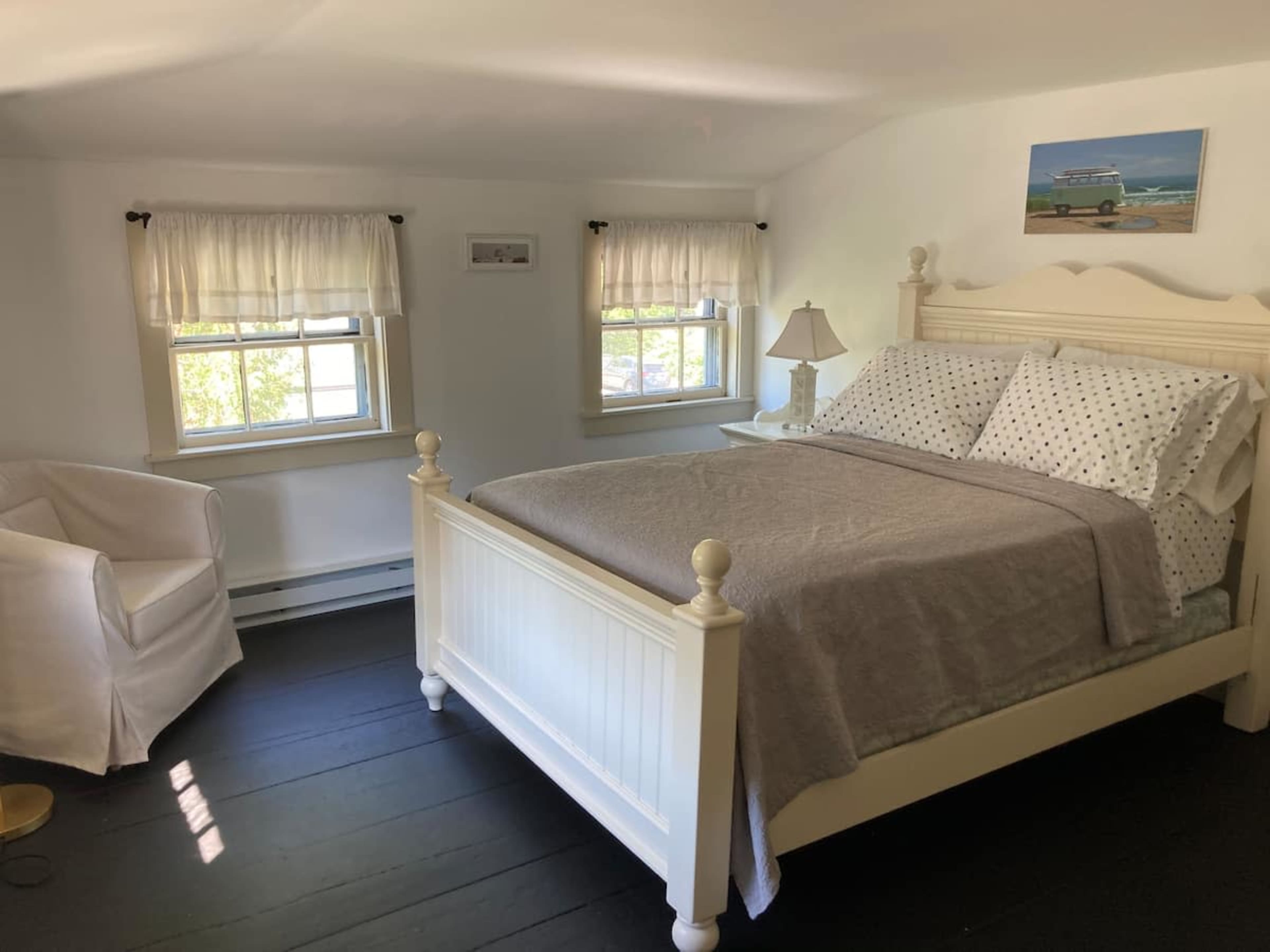A bright bedroom features a white bed with a gray blanket, two windows with sheer curtains, and a comfortable armchair in the corner.