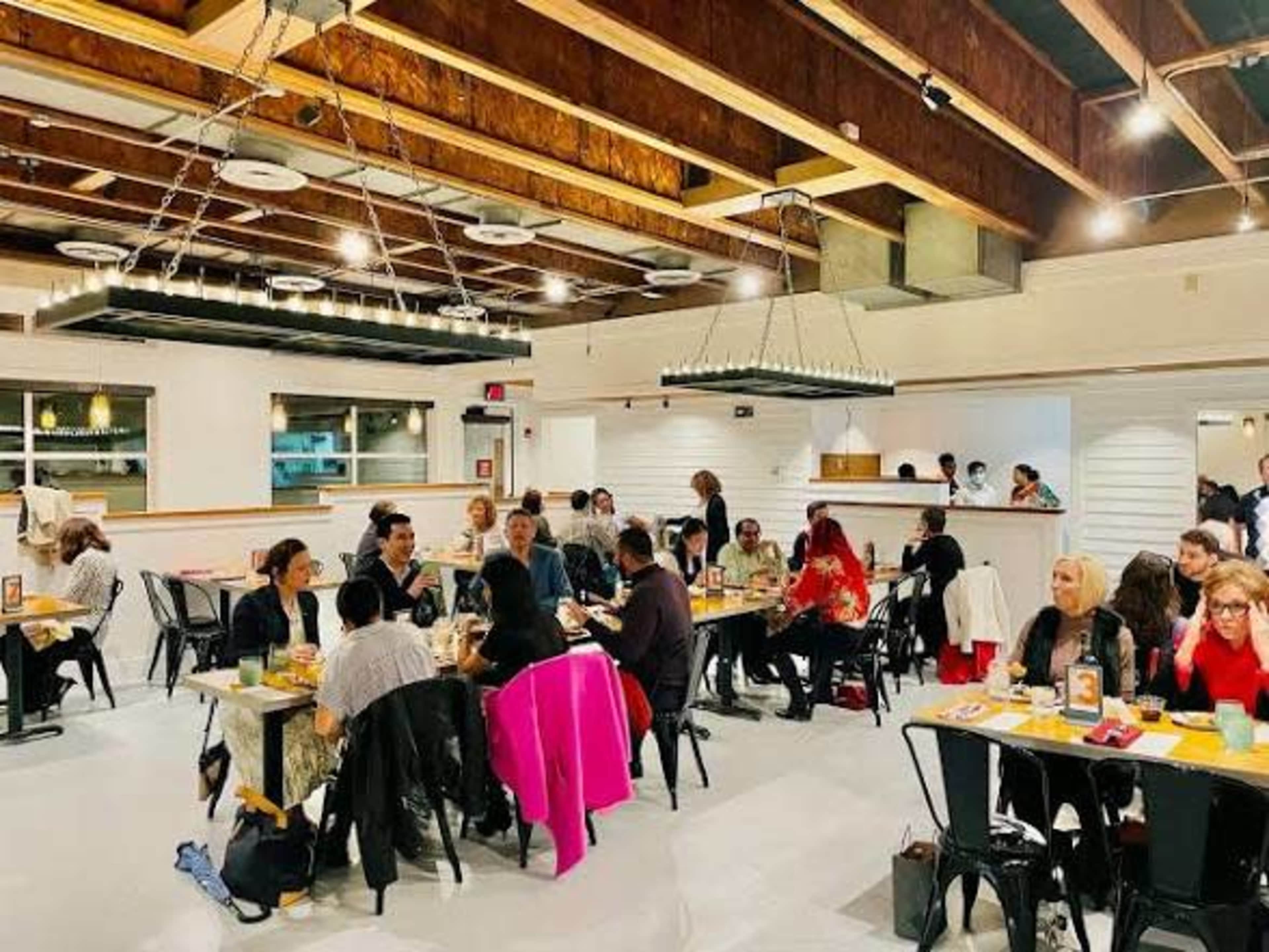 The image shows a modern restaurant interior with groups of people dining at various tables, featuring wood accents and bright lighting.