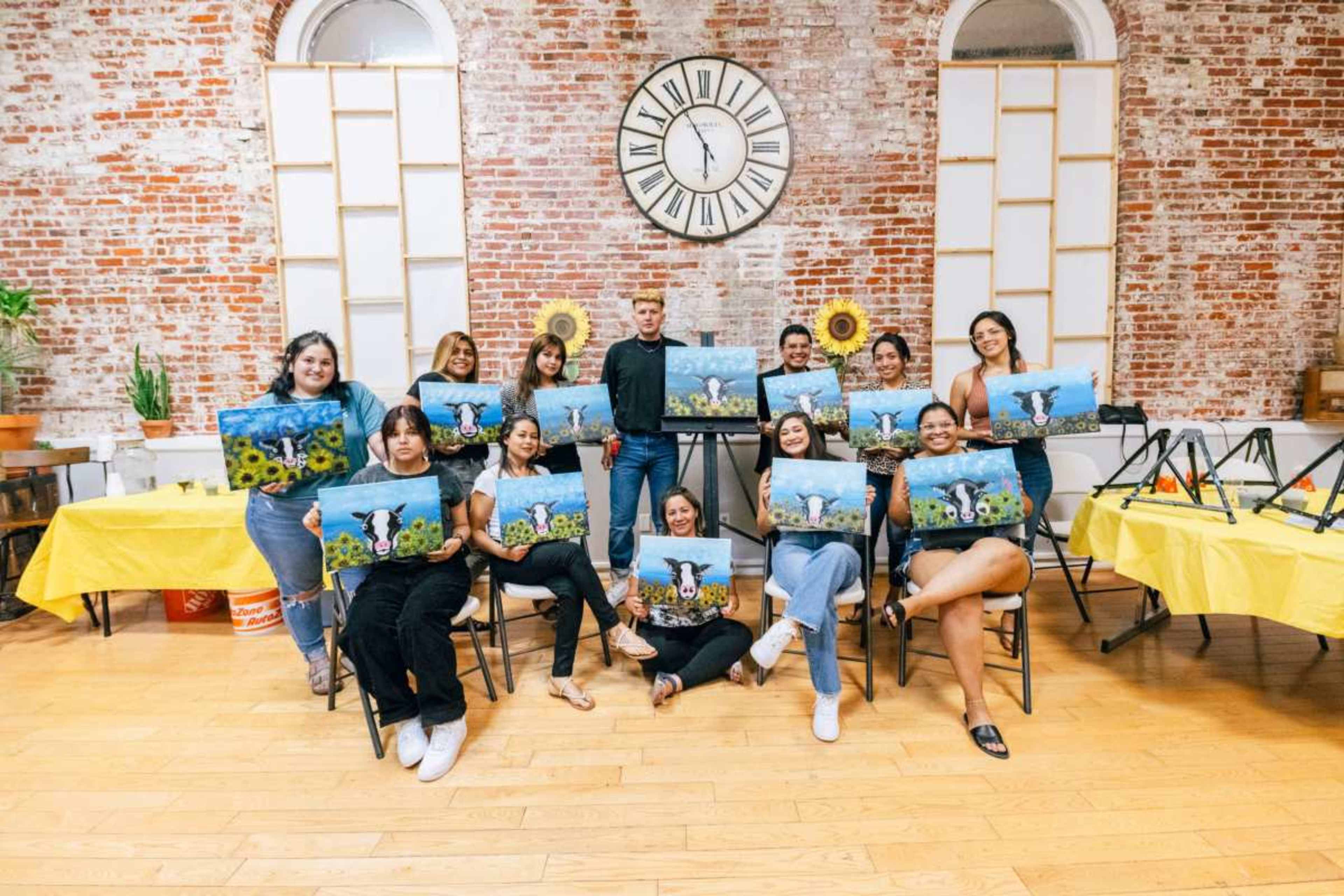 A group of people is posing with their completed paintings in a colorful art studio with a brick wall and large clock.