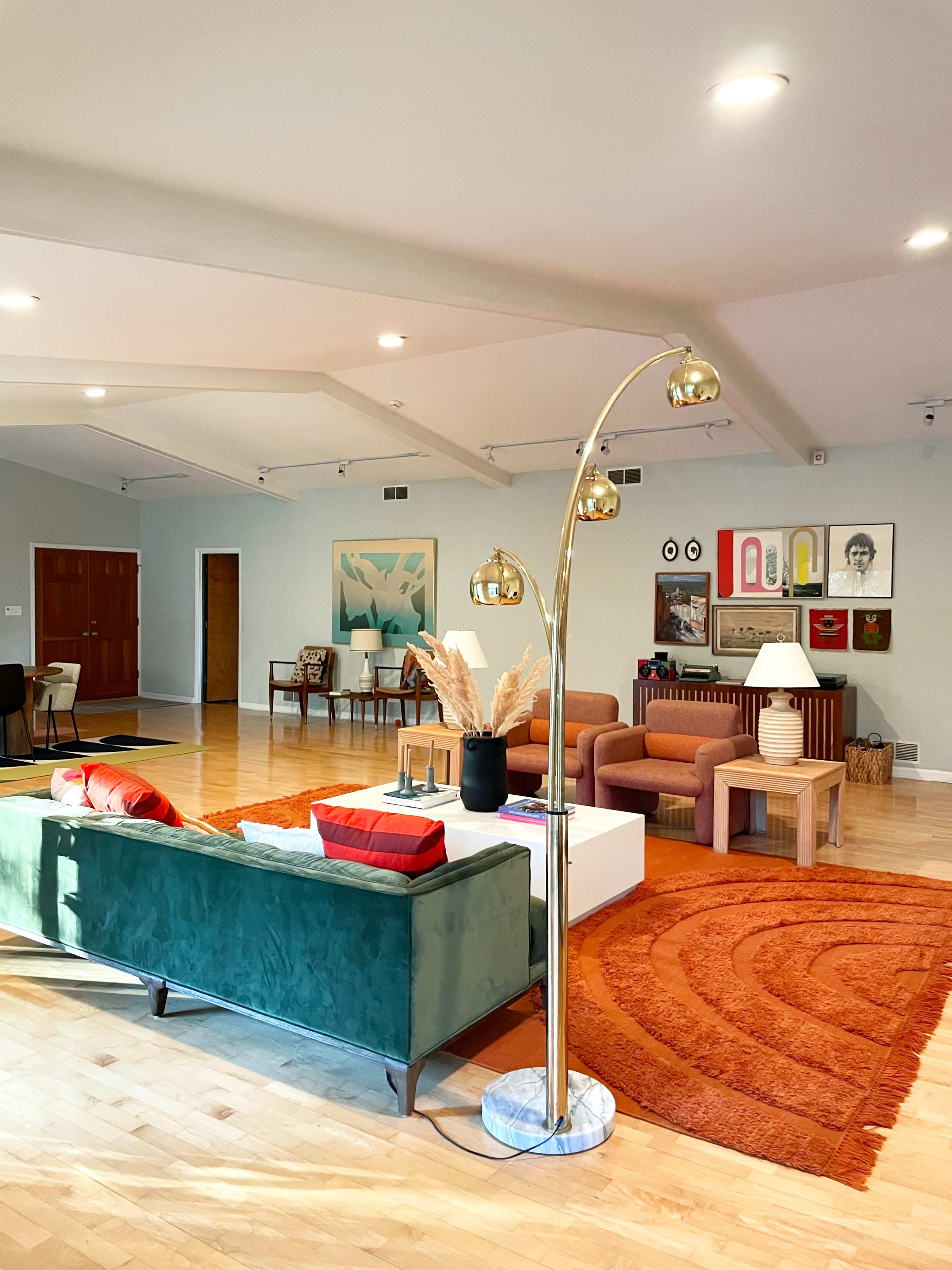 A spacious and brightly lit living room features a green velvet sofa, an arc floor lamp, a coffee table, and an orange rug.