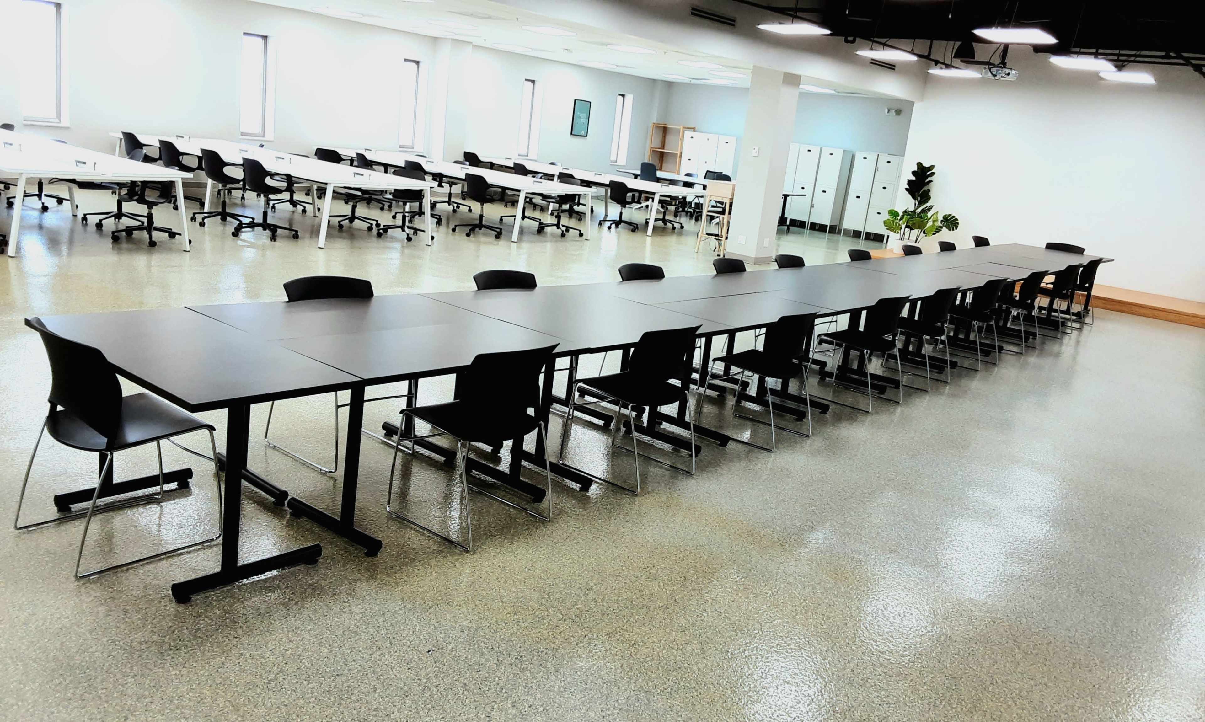 A long, rectangular table with black chairs is set up in a spacious room with polished concrete floors and numerous workstations in the background.