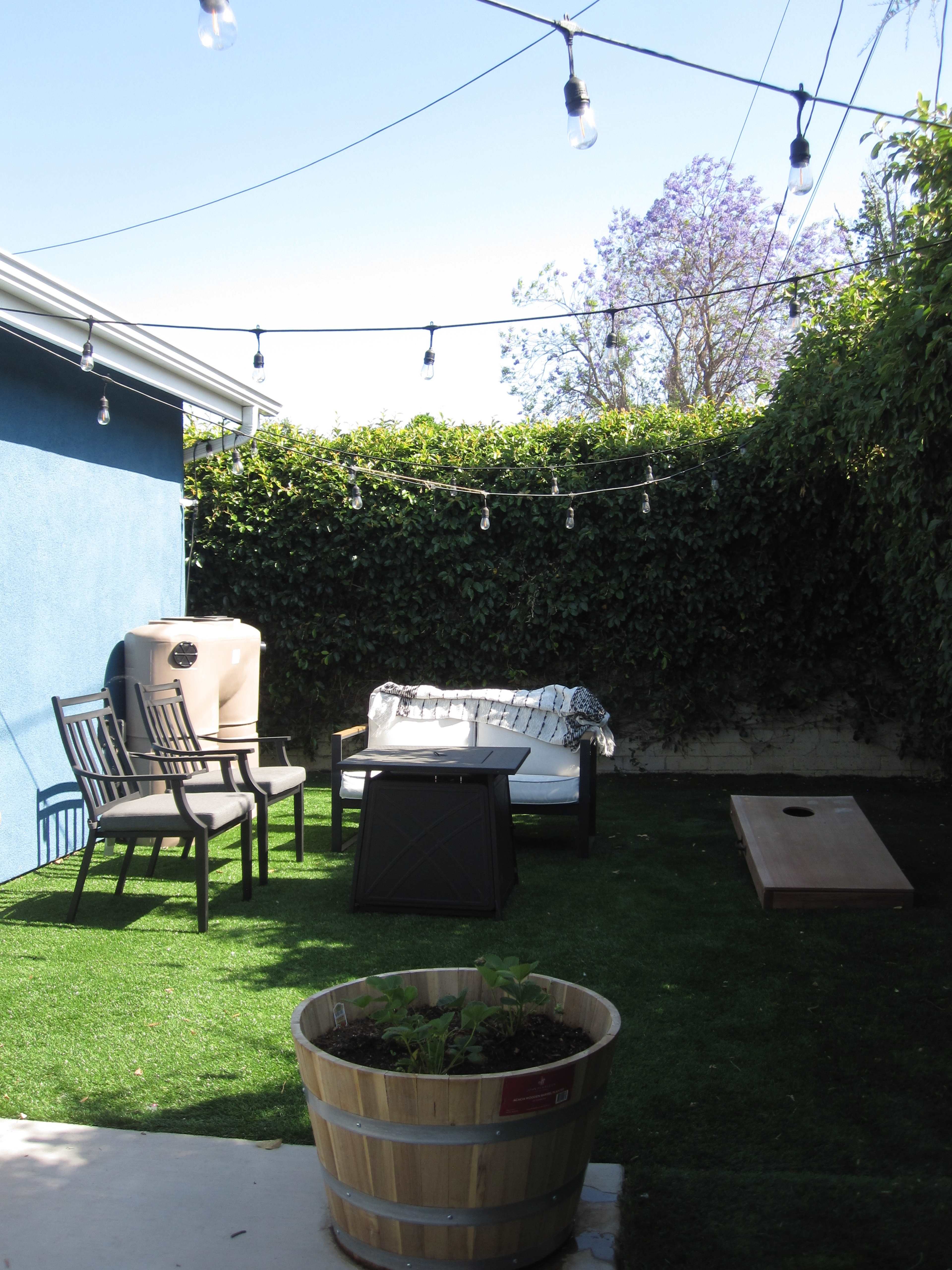 The image shows a small outdoor seating area with a couch, chairs, a coffee table, and a cornhole game surrounded by green plants and string lights.