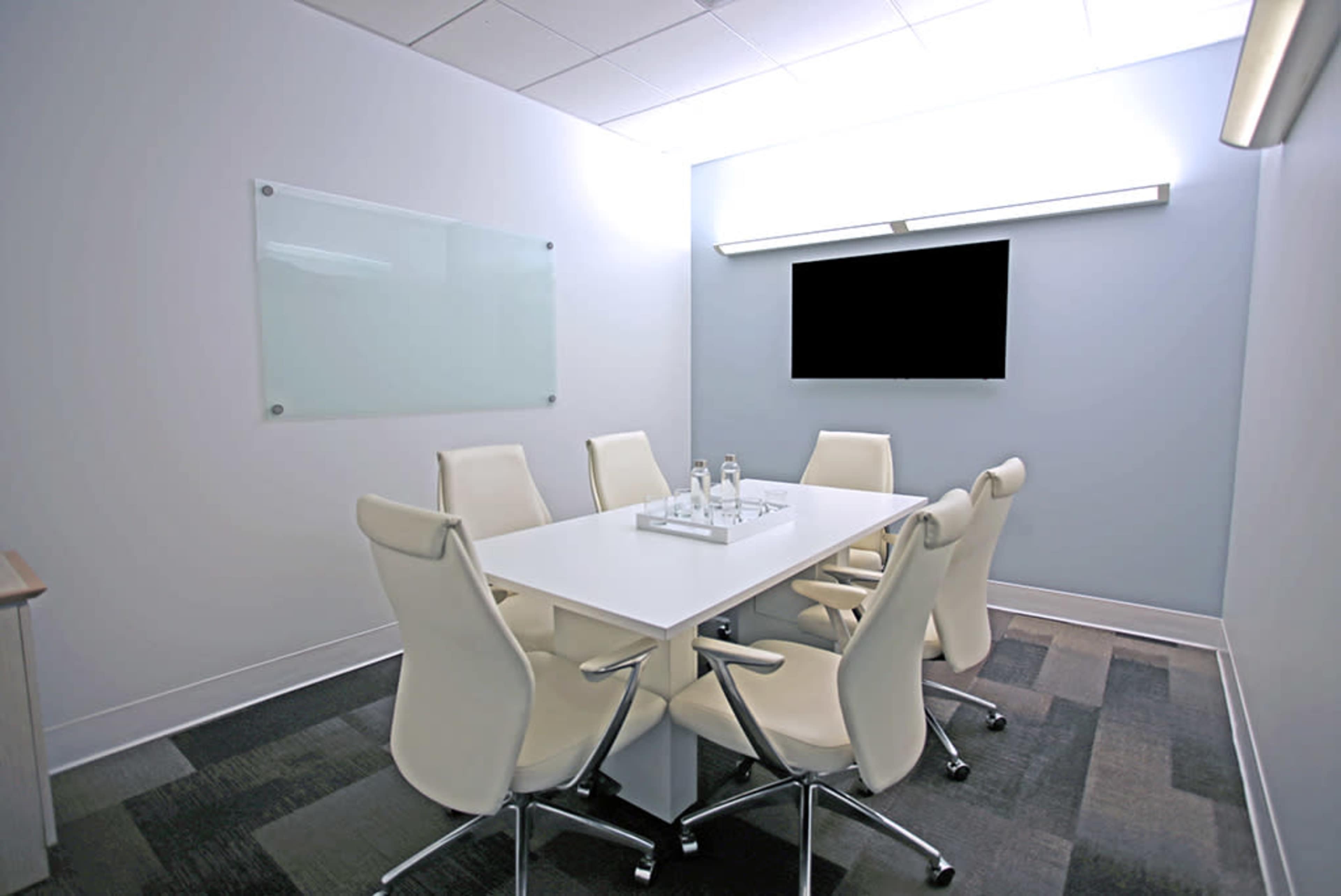 A modern conference room features a white table surrounded by six white chairs, with a glass whiteboard and a wall-mounted television.