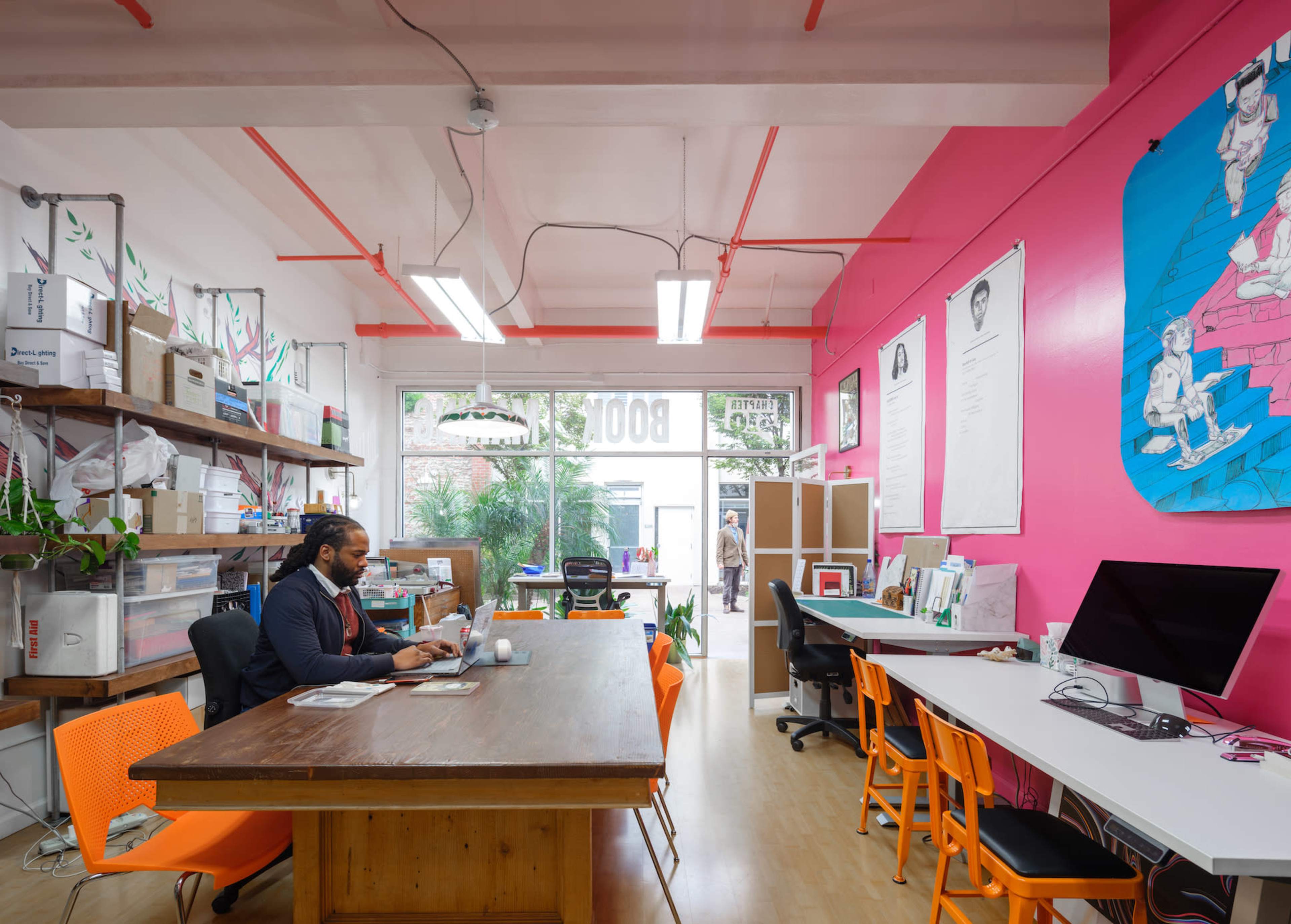 A man sits at a large wooden table in a brightly colored office space filled with plants, artwork, and desks.
