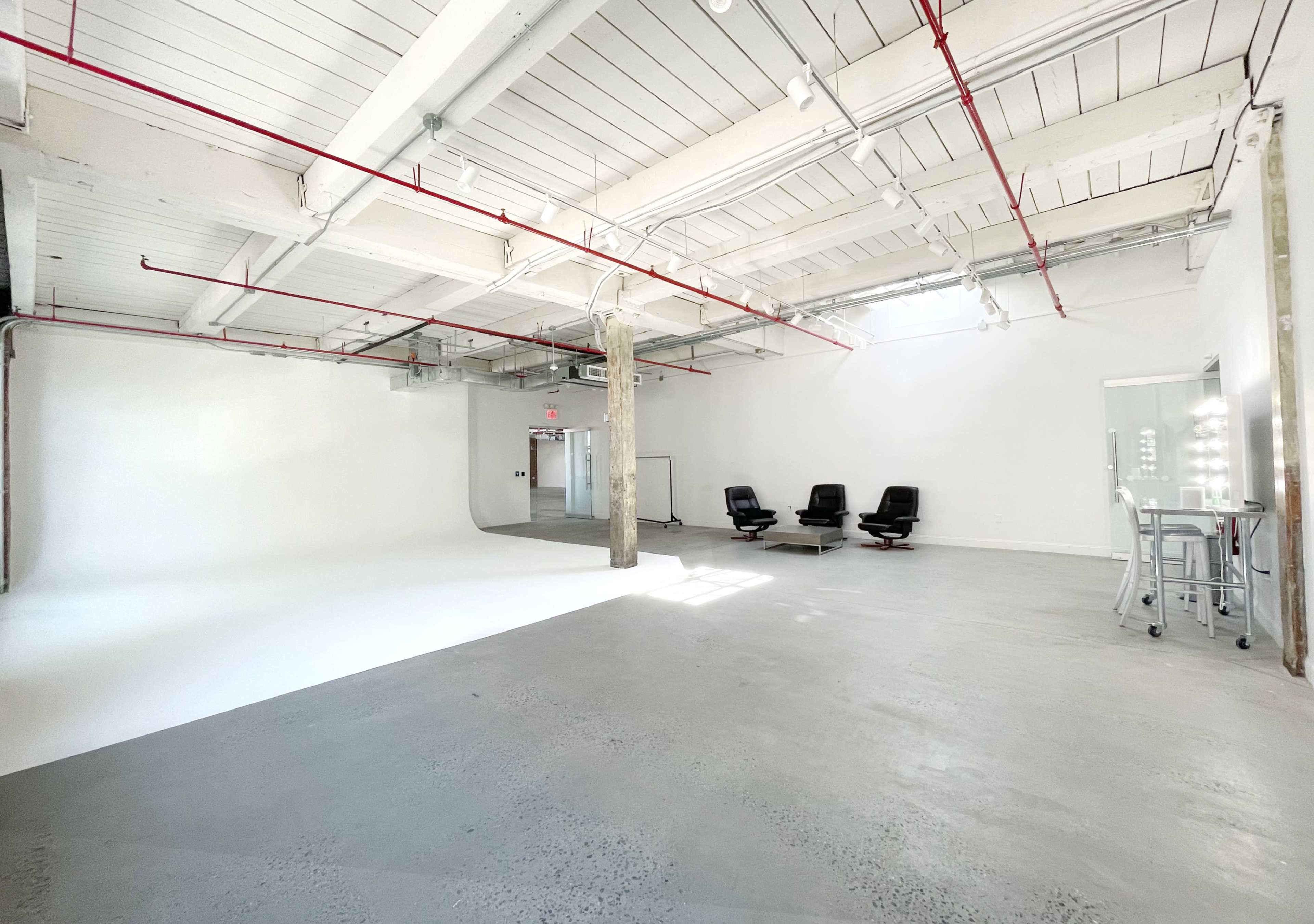 The image shows a spacious, empty studio with white walls, a concrete floor, and a large blank area for photography, accompanied by black chairs and a mirrored dressing table.