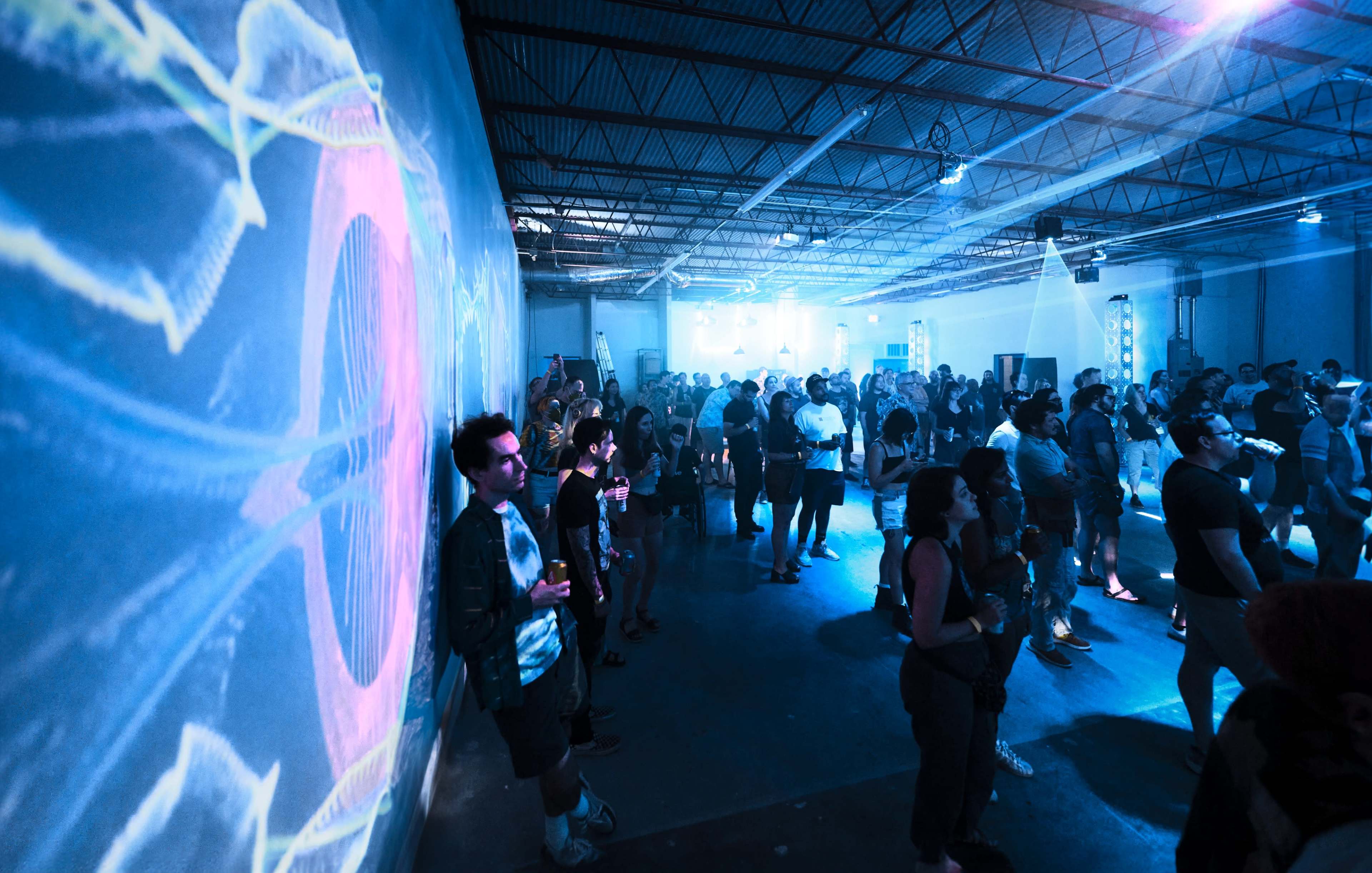 A crowd of people stands in a dimly lit warehouse, surrounded by colorful, swirling light projections on the walls.