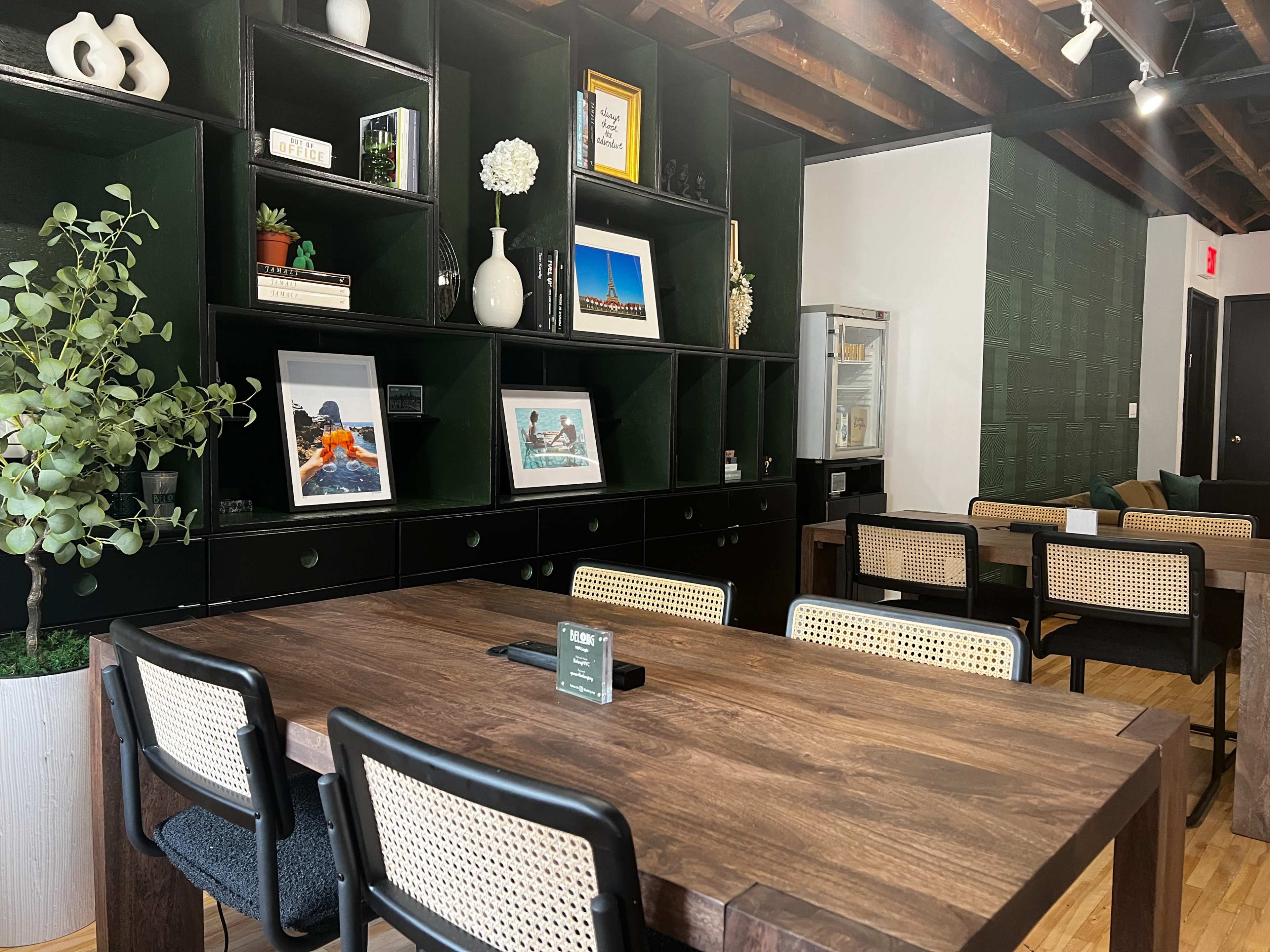 The image shows a modern, minimalist interior space with wooden tables, black shelving displaying decorative items, and plants.
