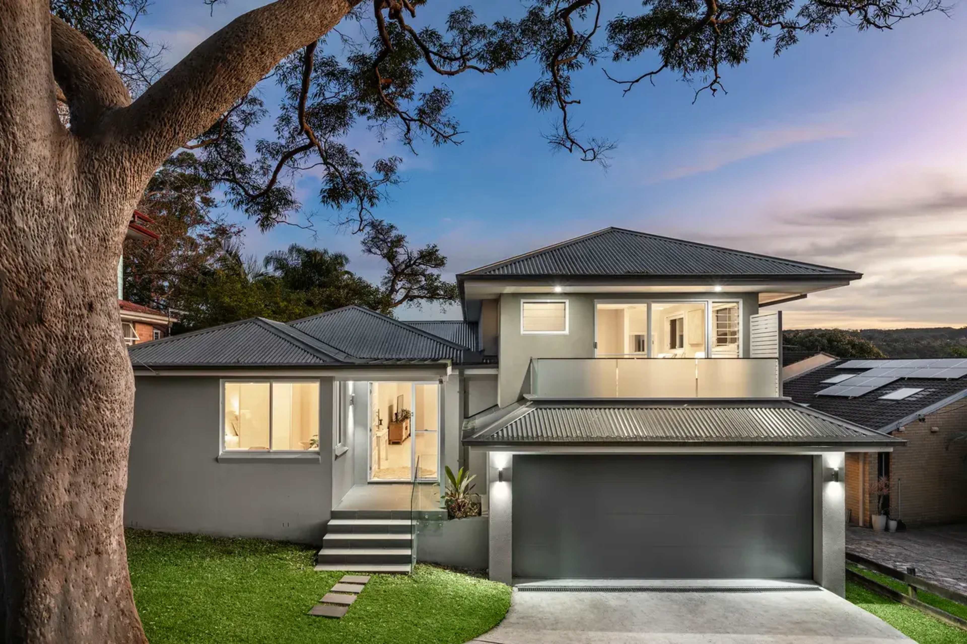 A modern two-story house with a grey exterior and a garage sits under a large tree in the evening light.