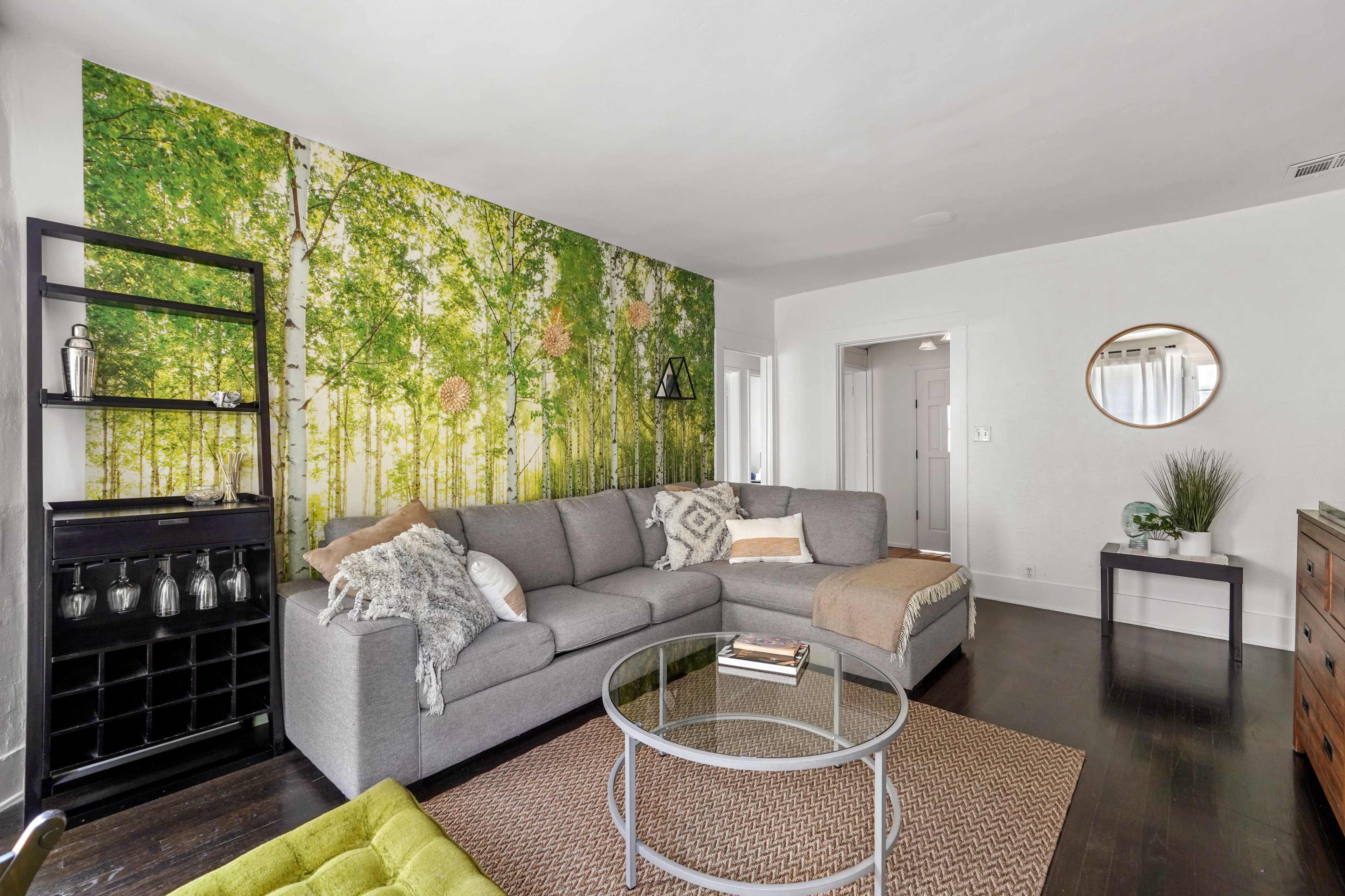 The living room features a gray sectional sofa, a glass coffee table, and a nature-themed mural of trees on one wall.