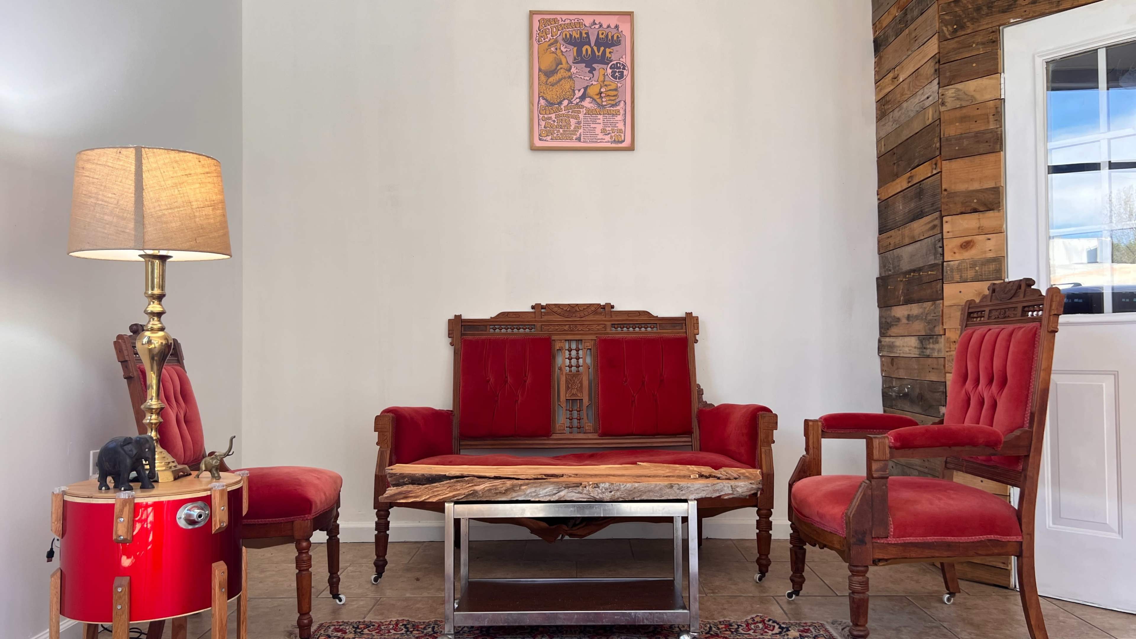 The image shows a cozy room with red upholstered seating, a wooden coffee table, and a lamp beside a small drum on the side.