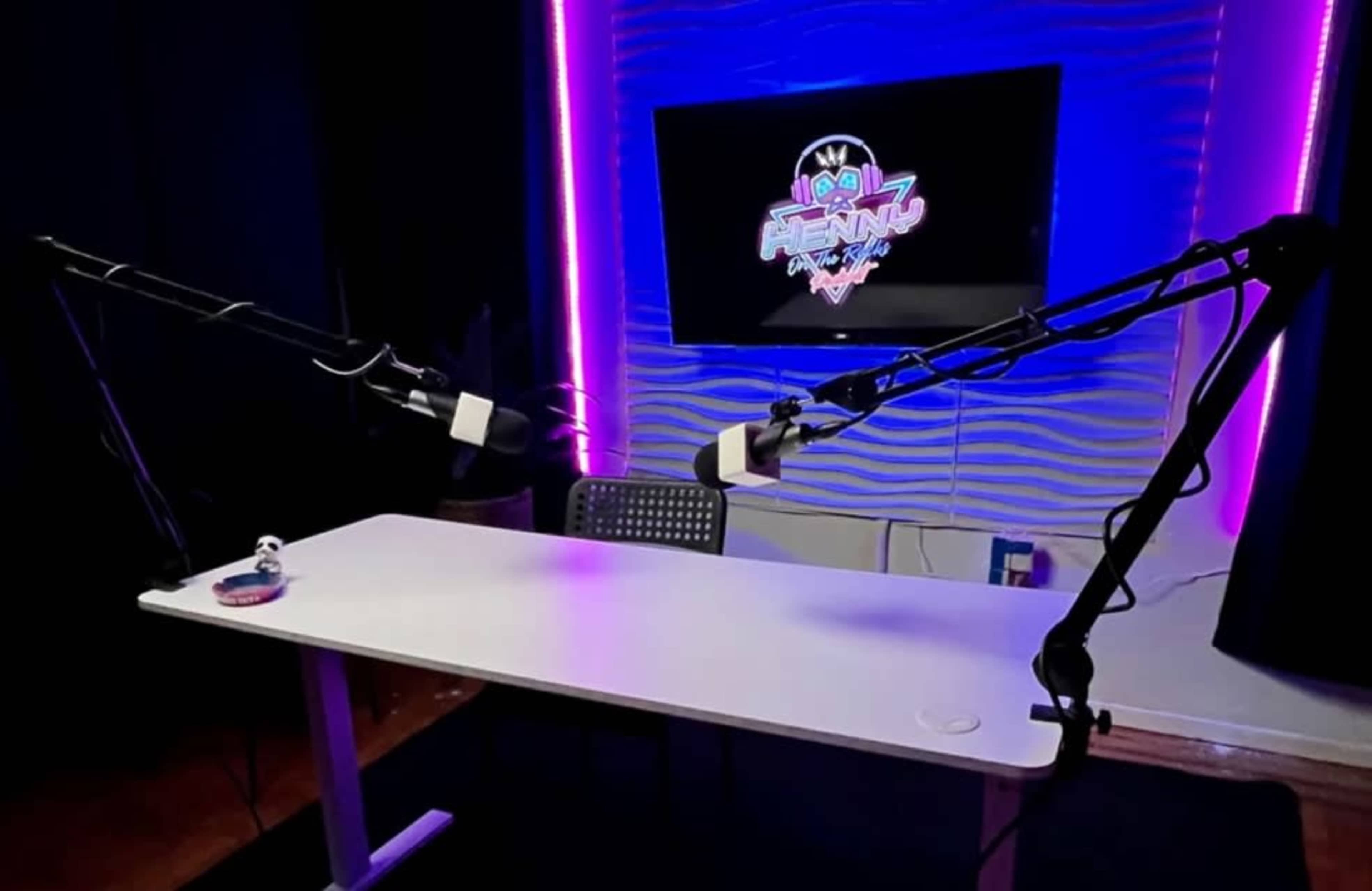 The image shows a podcasting studio setup with a white table, two adjustable microphone arms, and a screen displaying a colorful logo in the background.