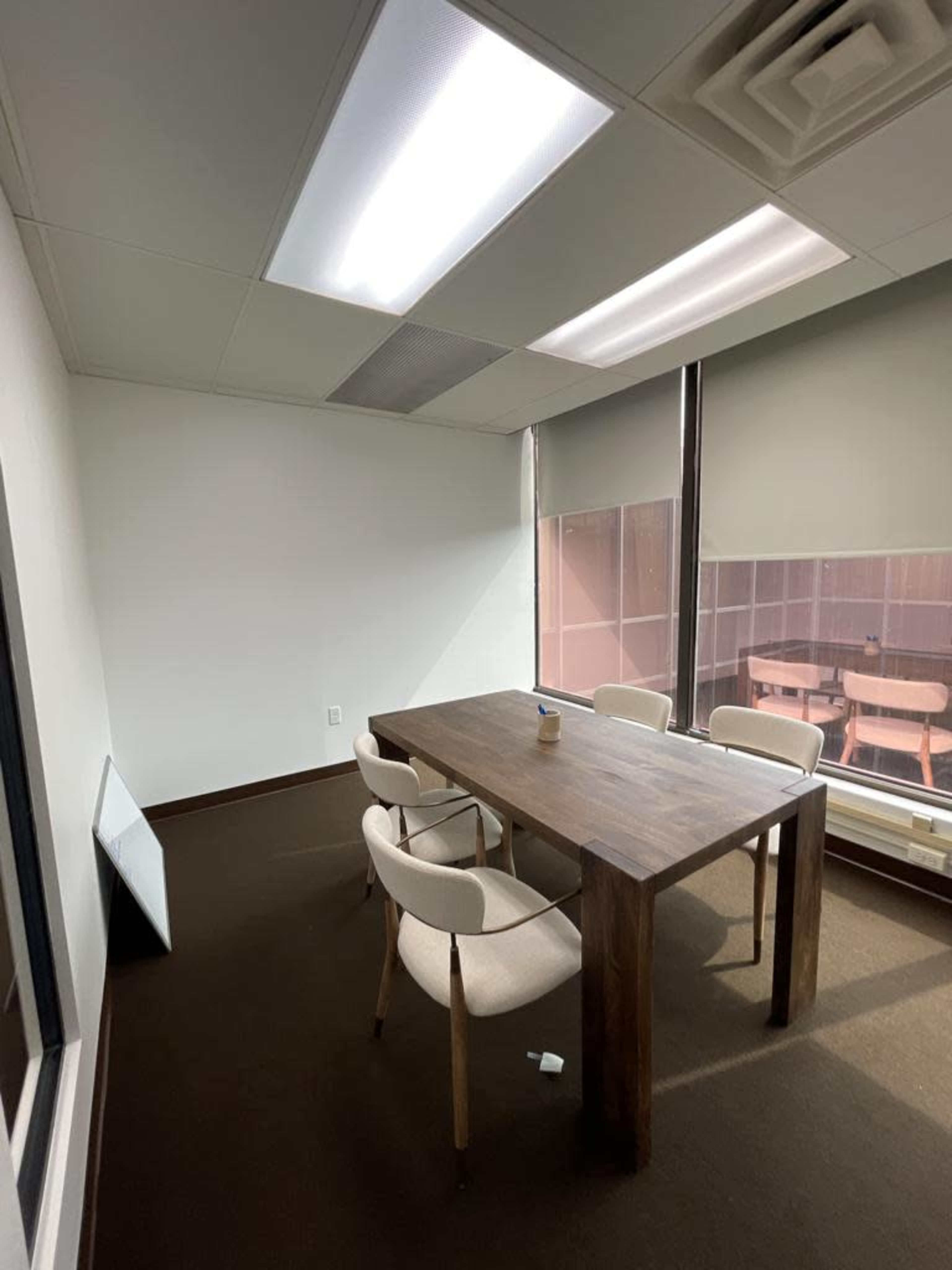 A small meeting room with a wooden table surrounded by four chairs, adjacent to a window with a view of another room.