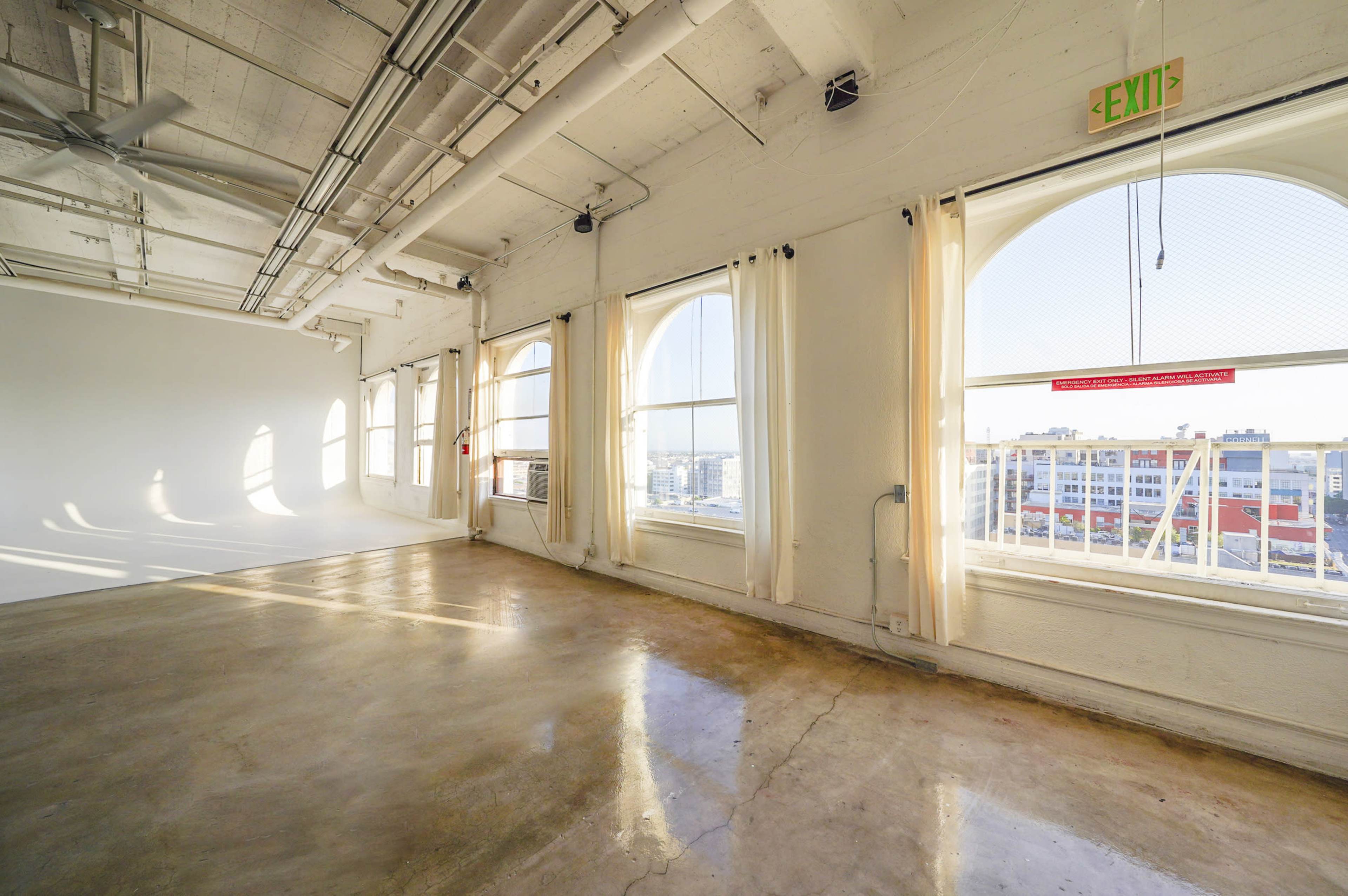 The image shows a spacious, brightly lit room with large windows, white walls, and a concrete floor.